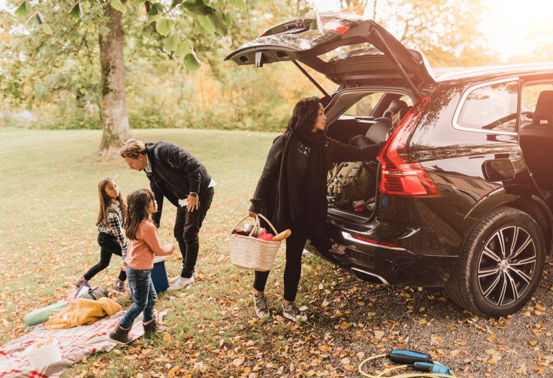 Family of four getting ready for a picnic, next to a black car at a park.