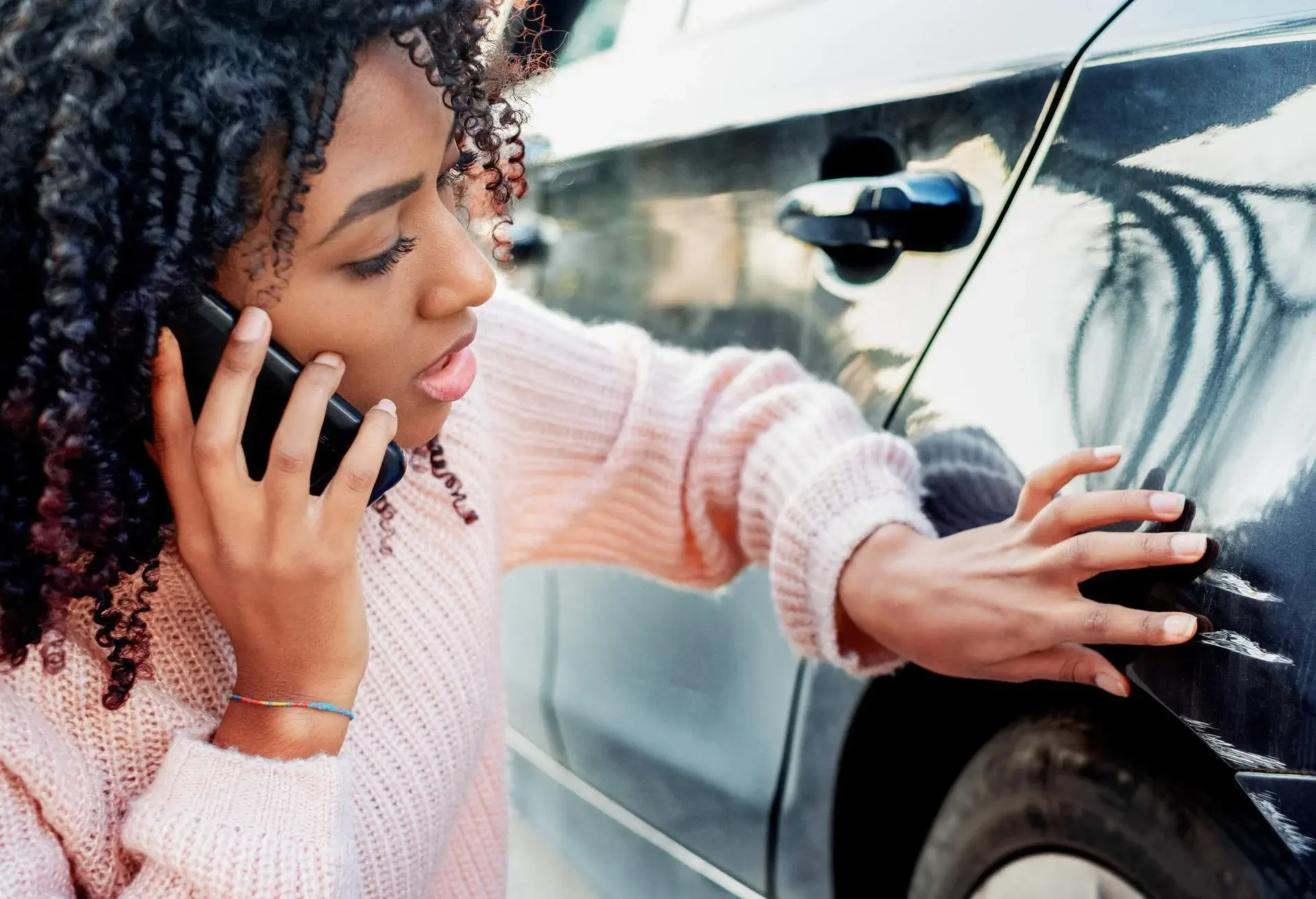 A woman with curly brown hair is on the phone while looking at some damages on a car