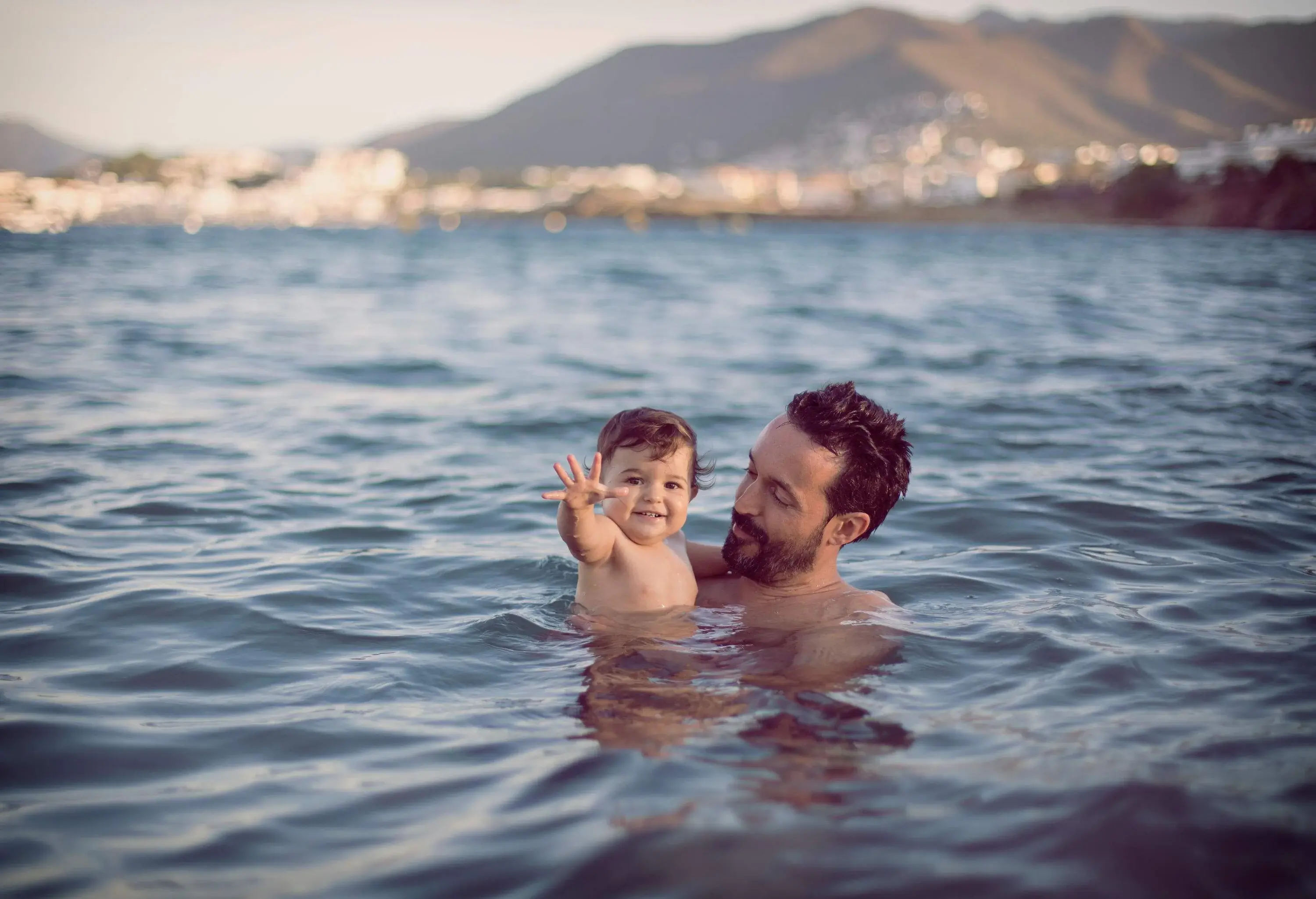 A bearded man carries a child while immersed in the ocean.