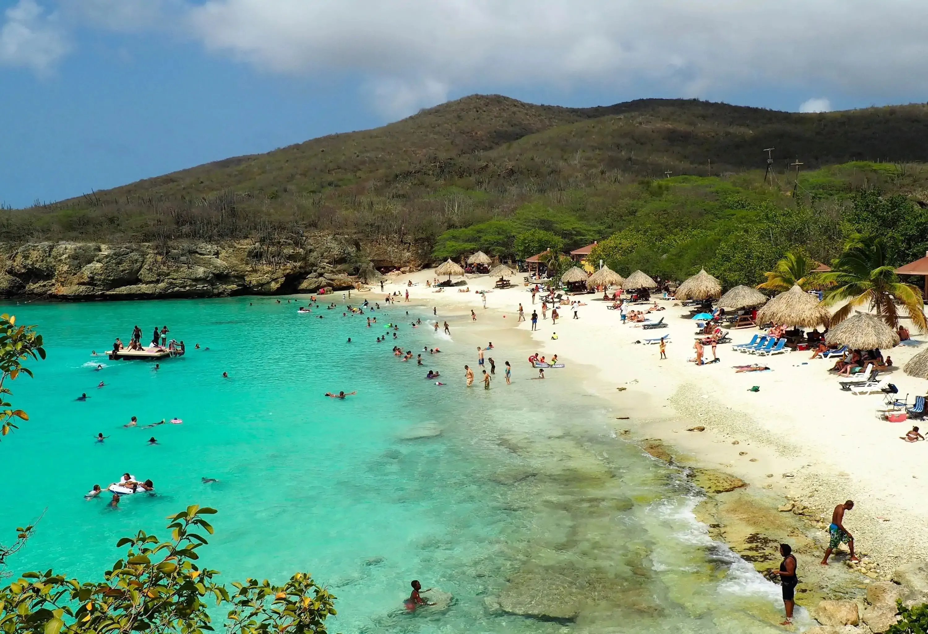 A beach with beachgoers enjoying the sun and waves, rustic thatched huts providing shade, and lush vegetated slopes serving as a picturesque backdrop.