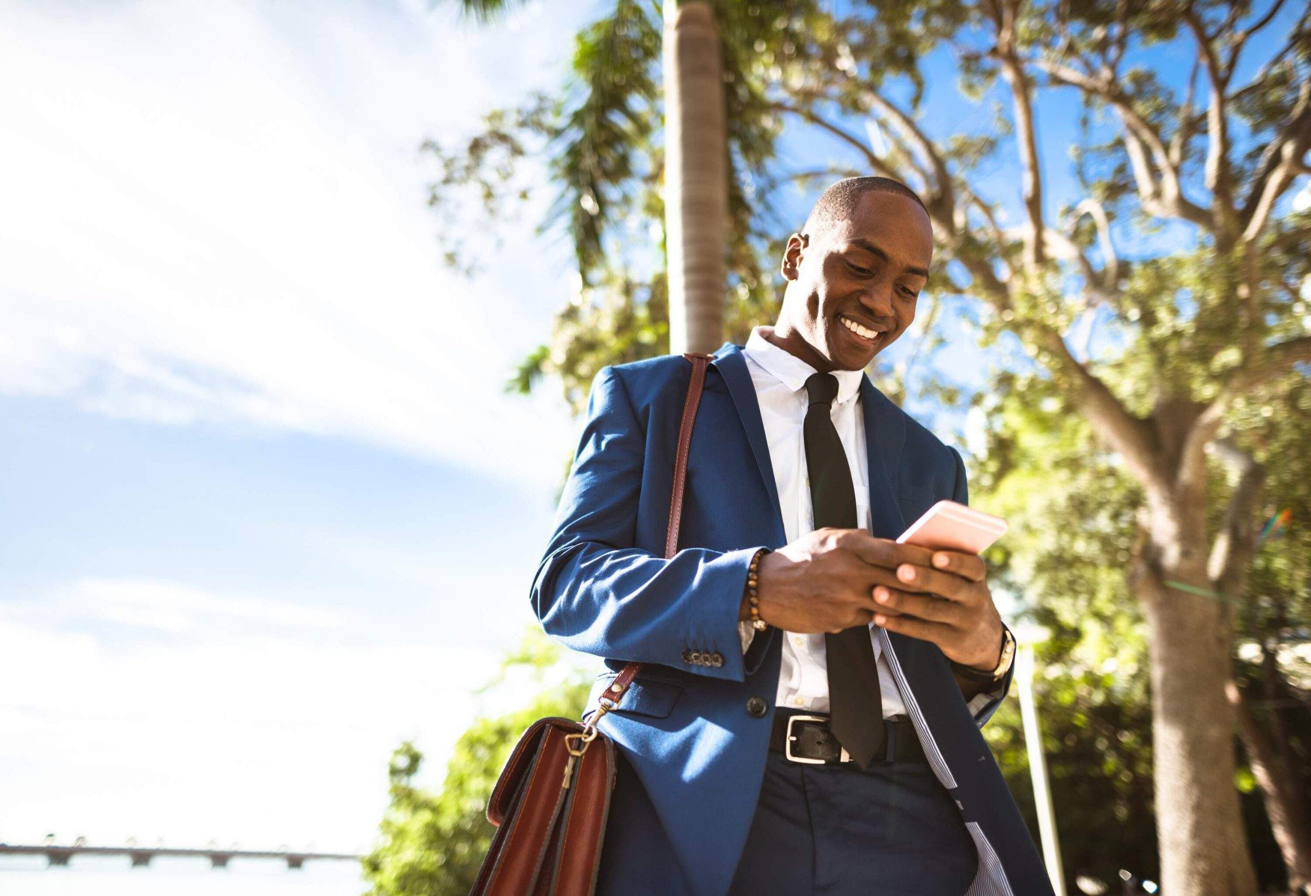 A businessman with a brown leather bag slung over his shoulder smiling as he checks on his phone.