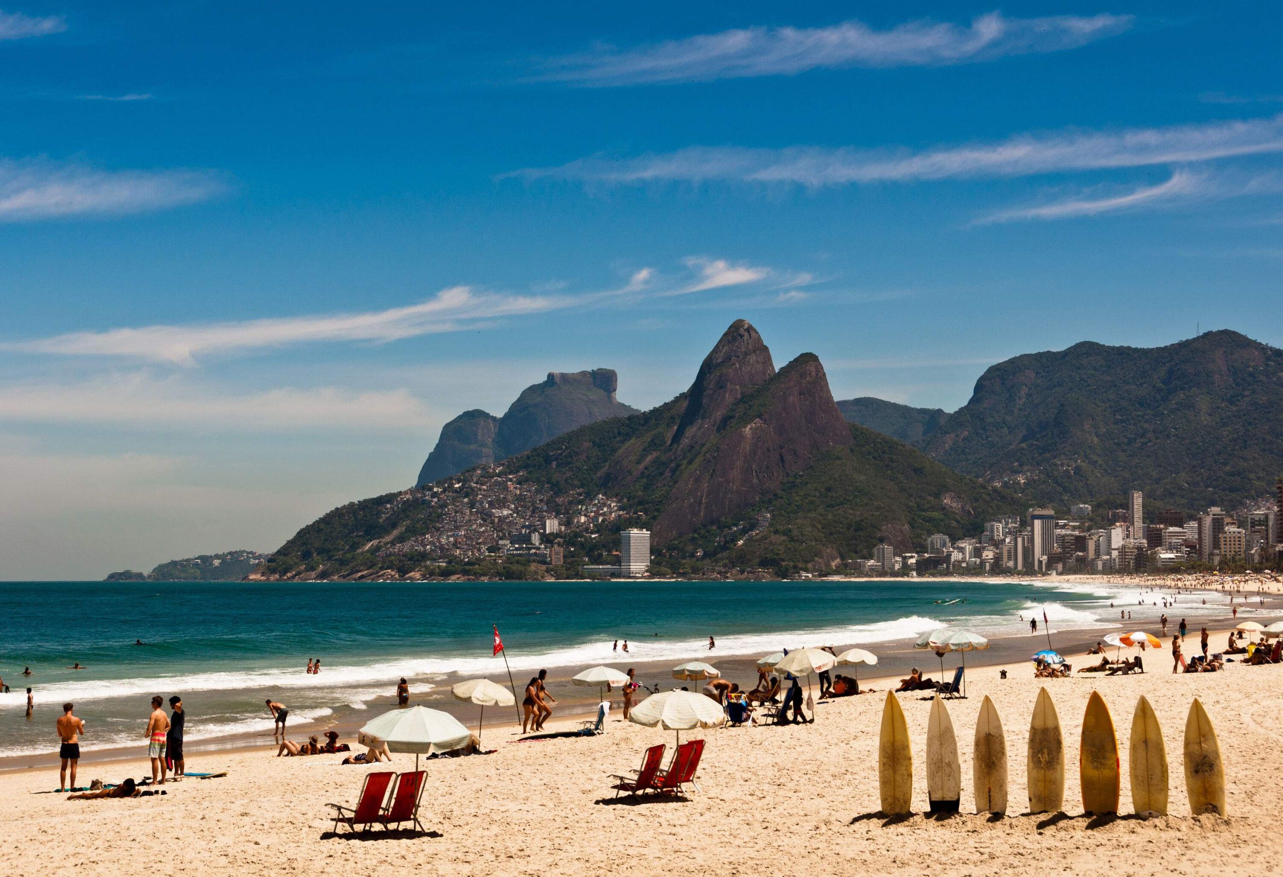 A populated beach with surfboards erected on the sand and umbrellas strewn about as people bask in the sun and bathe in the sea.