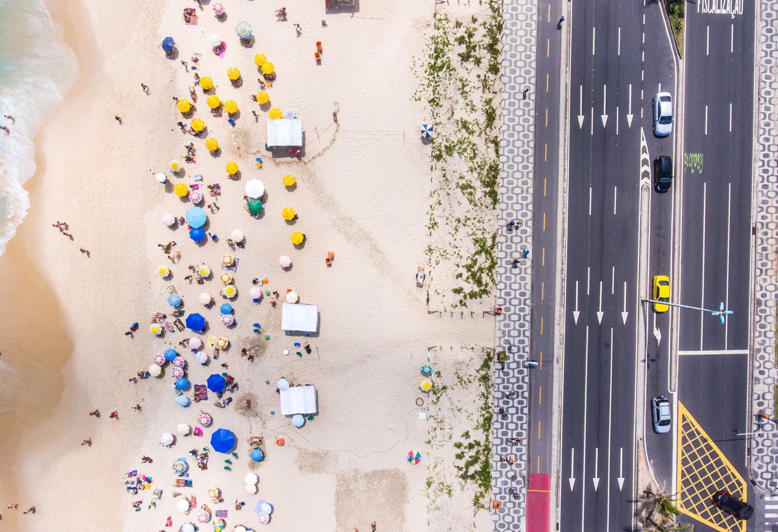An aerial top view of a beach with a sandy shore dotted with beachgoers and colourful parasols stretches alongside the road, creating a vibrant and lively coastal scene.