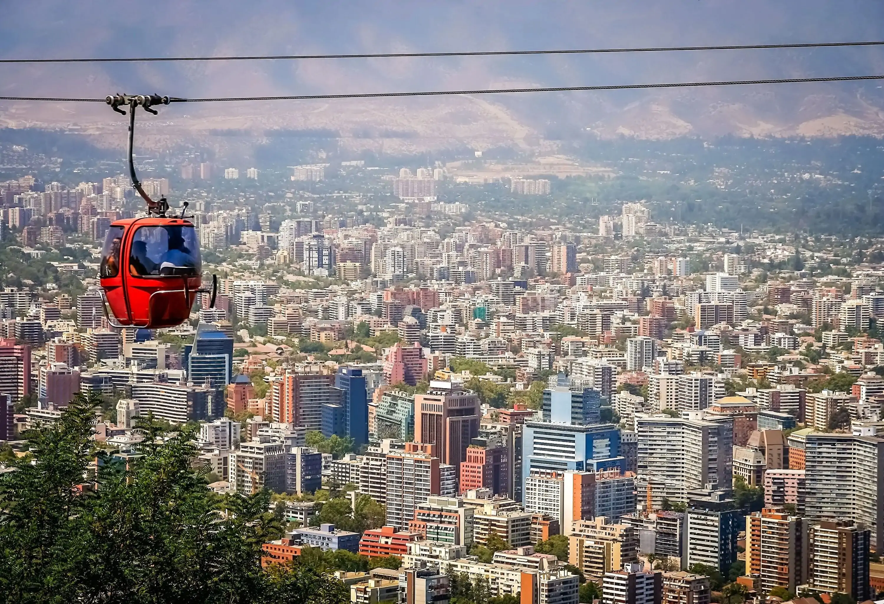 An orange cable car travelling on a cable with views of a cluster of high-rise city buildings.