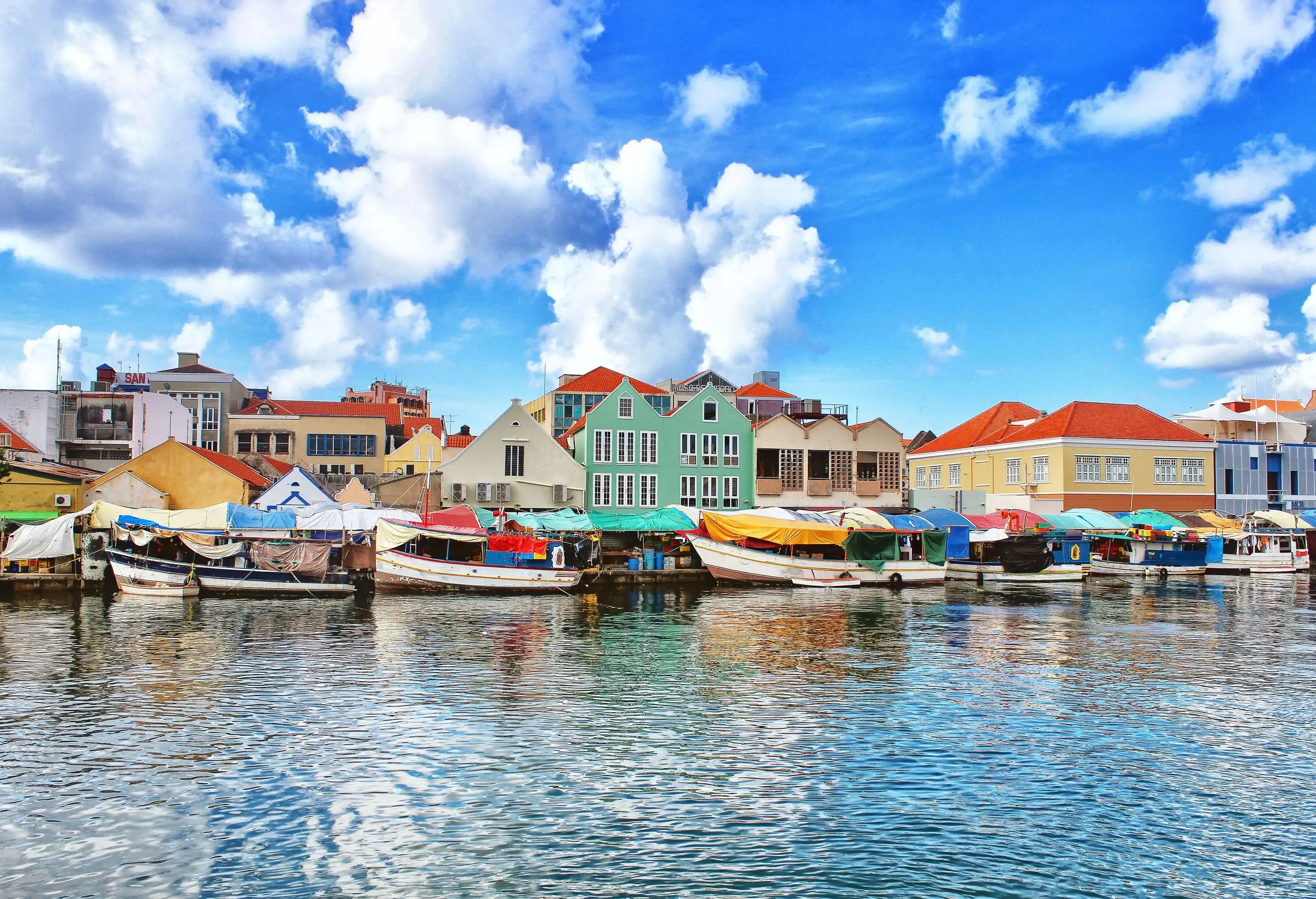 A lively promenade adorned with colourful boats, brimming with merchandise, gracefully moors along the waterfront, while buildings provide a charming backdrop.