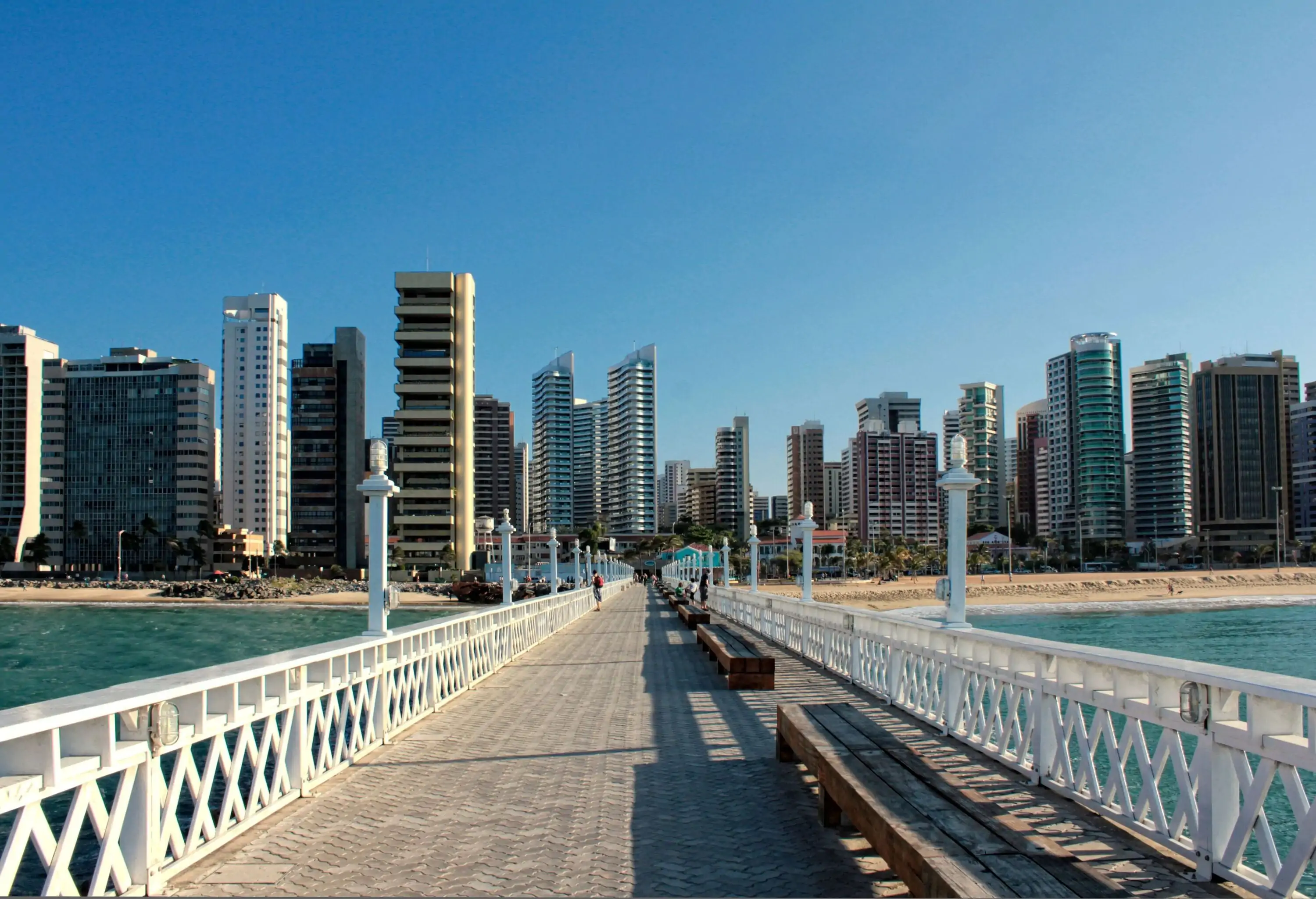 A picturesque pier with white guardrails stretching ahead towards the sandy beach forms a stunning and vibrant backdrop.