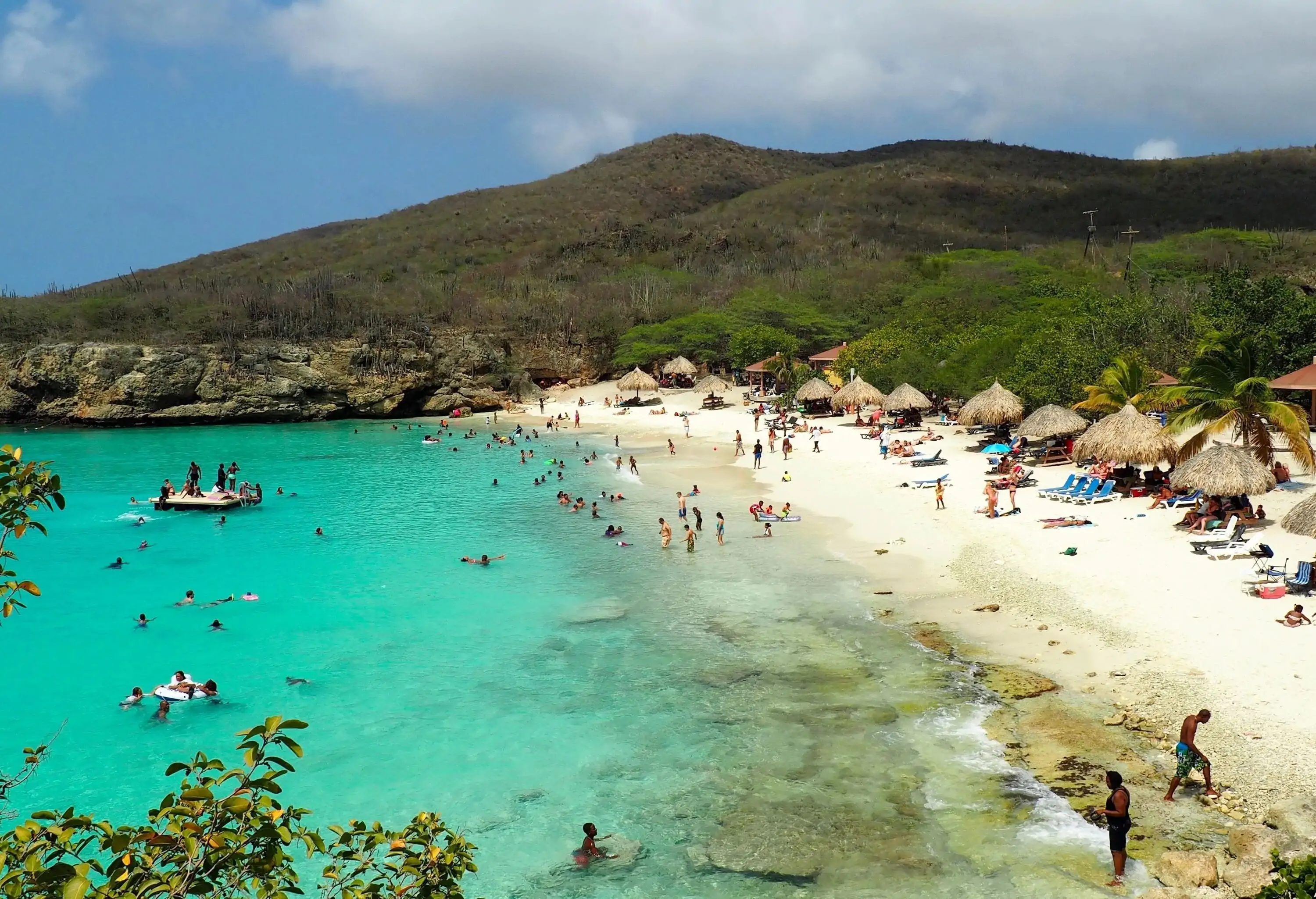 A beach with beachgoers enjoying the sun and waves, rustic thatched huts providing shade, and lush vegetated slopes serving as a picturesque backdrop.