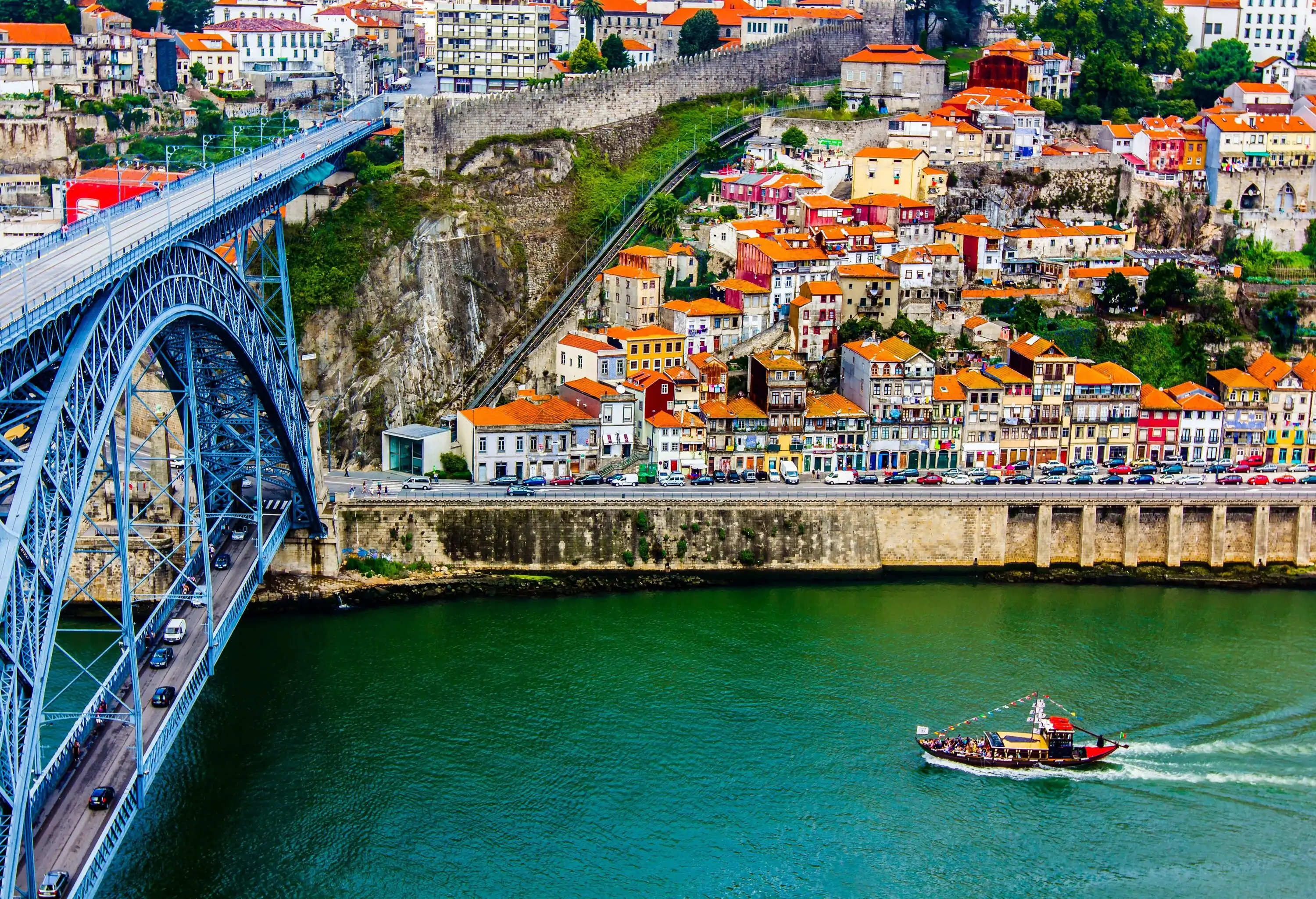 A boat loaded with people sails on a tranquil river passing through a metal arch bridge along a hill with compact houses.