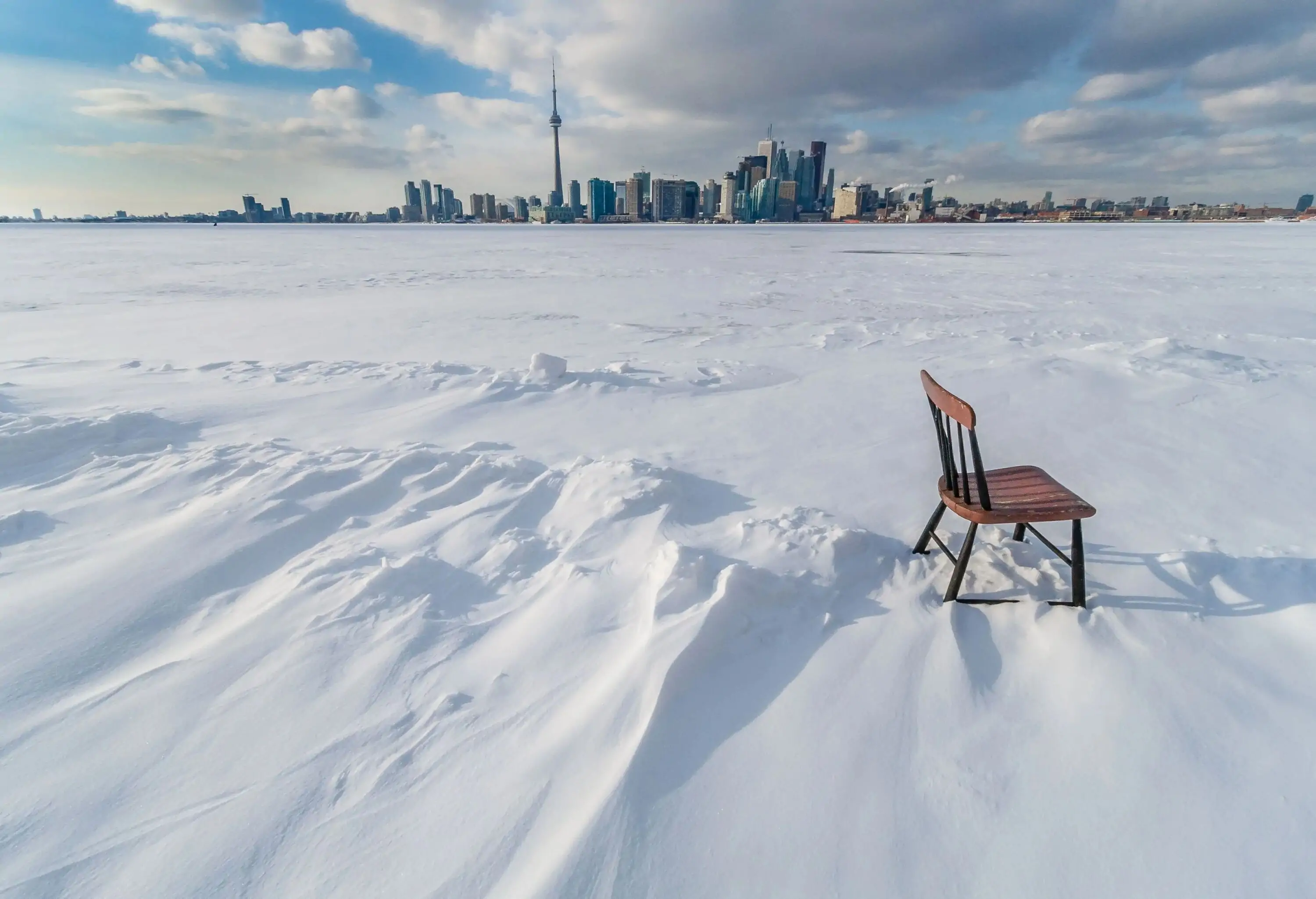 A lone wooden chair on a thick blanket of snow with high-rise structures in the background.