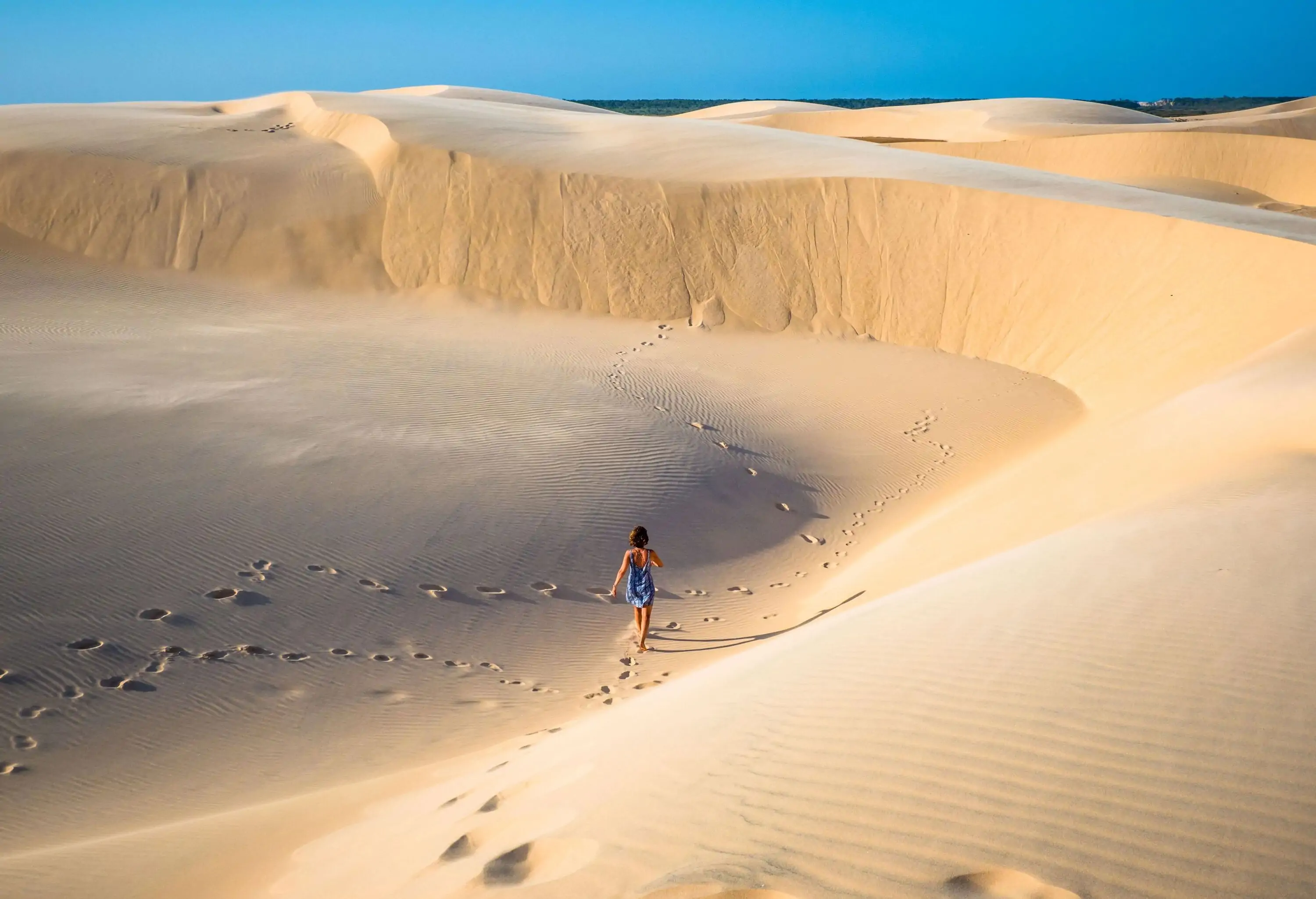 A woman wandering across a fine sand dune with traces of footprints behind her.