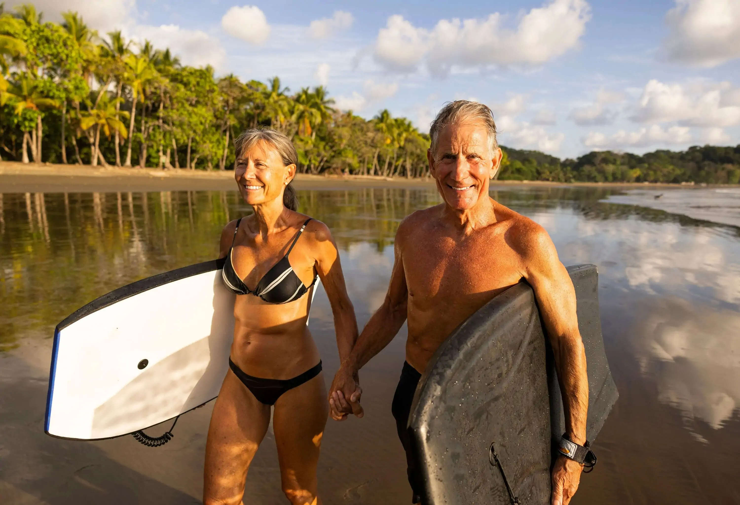 Senior couple carrying SUPs coming out of water on a tropical beach at golden hour