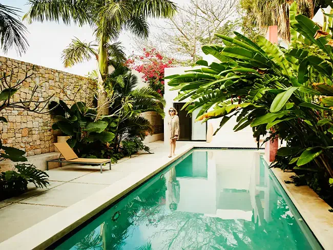 Foto de uma piscina em um ambiente que parece uma casa, cheia de árvores e plantas verdes. É possível ver um muro de pedras no lado esquredo, e ao fundo, uma mulher caminha em direção à câmera.