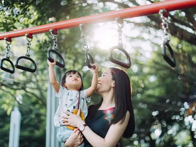 foto de uma mãe segurando o filho no colo, que está esticando os braços para se pendurar em um brinquedo de praça