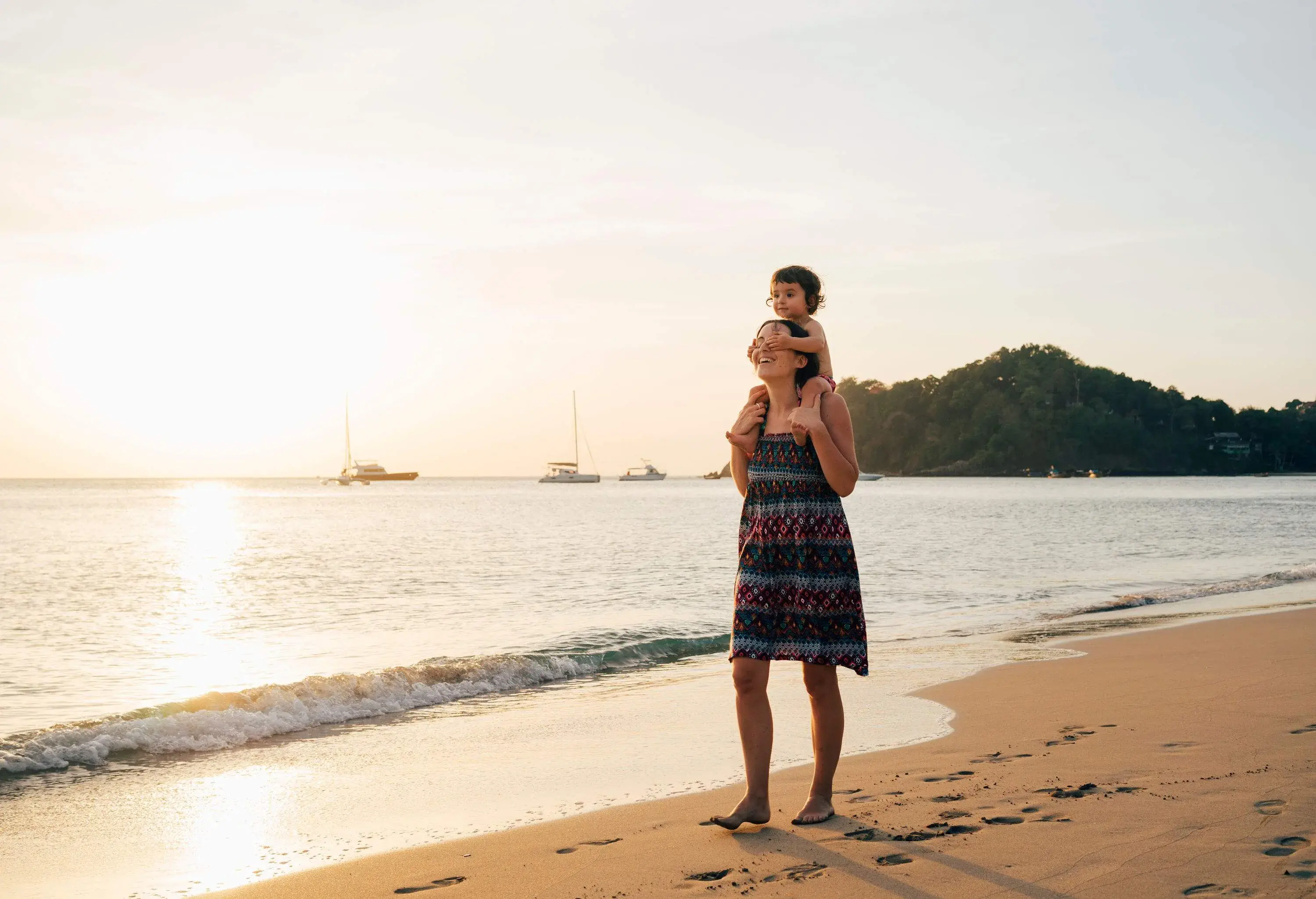 A mother with her daughter on her shoulder walks on the beach at sunset.
