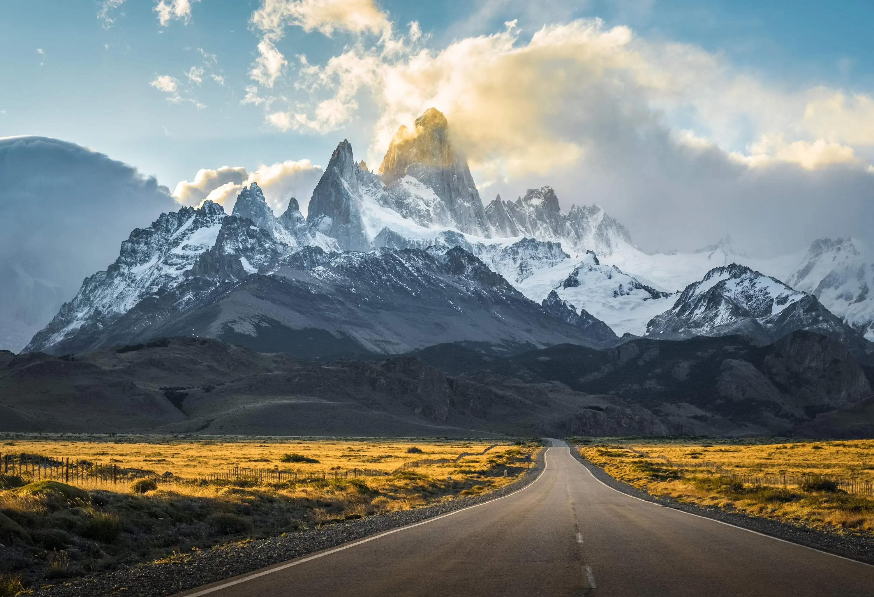 A straight road running across a grass field backed by a serrated mountain with snowy peaks.