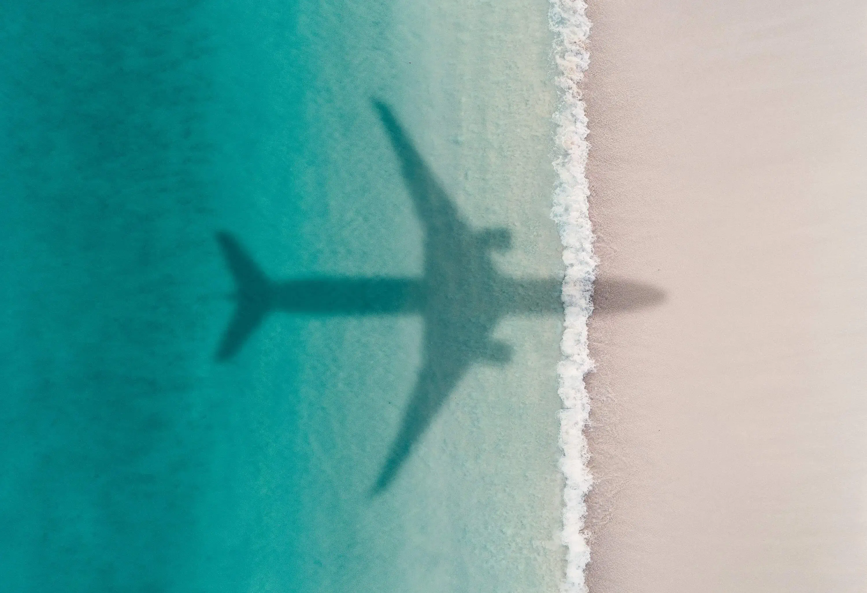 An airplane's shadow casts on a beach with turquoise waters.