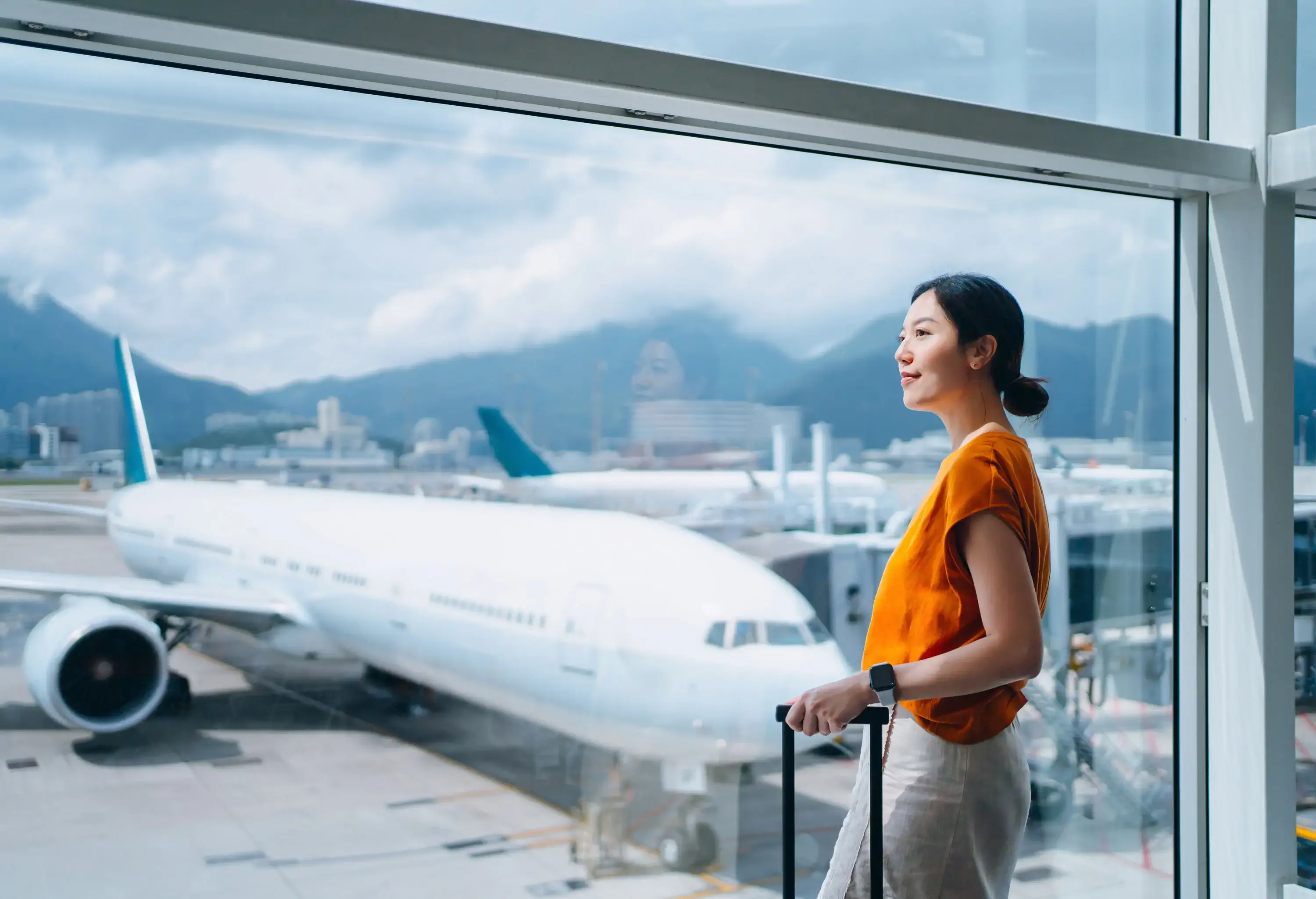 Young woman carrying suitcase, looking through window at airport terminal while waiting for departure. Ready for a trip. Business travel. Travel and vacation concept