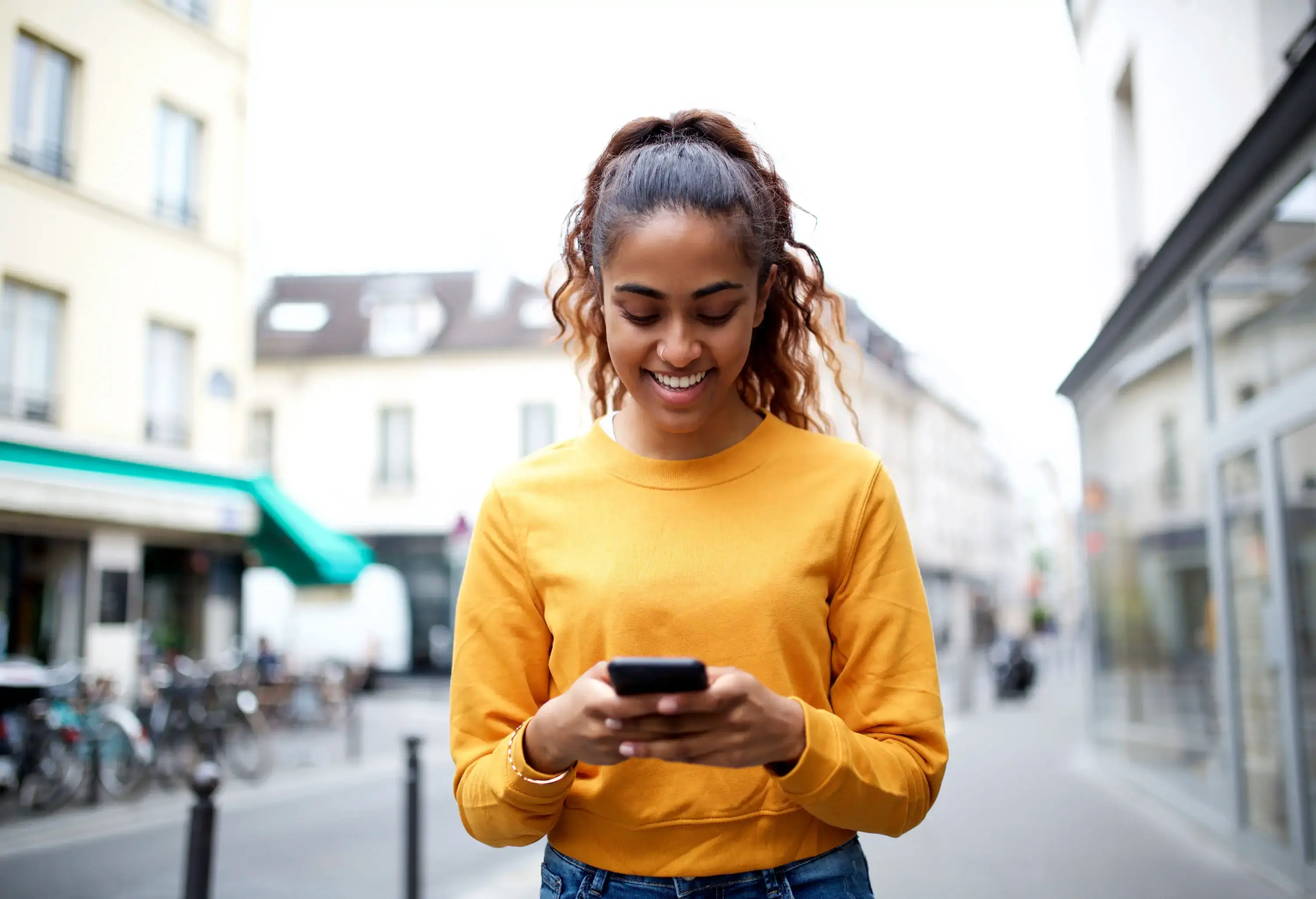 A smiling young lady uses her mobile phone while walking on the street.