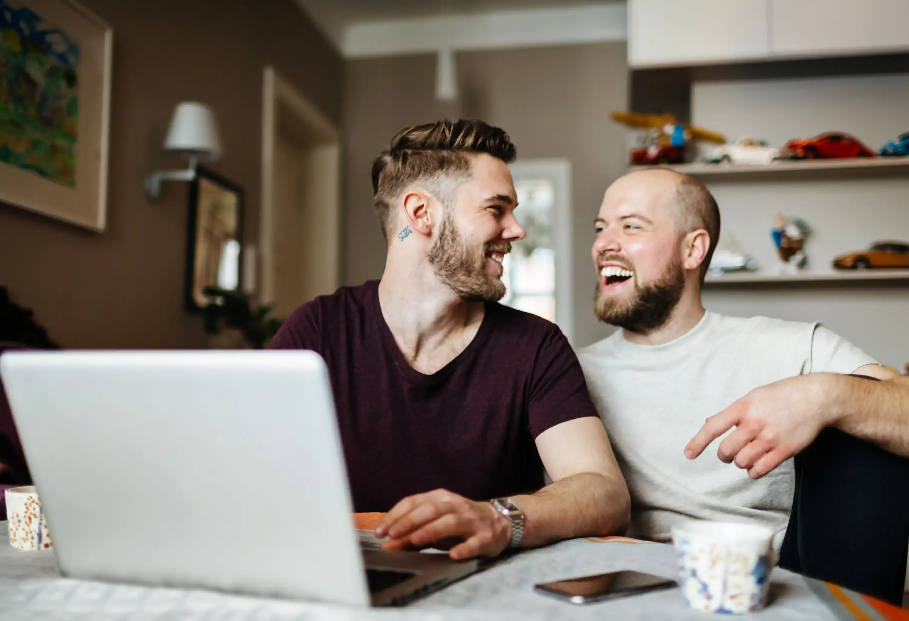 A happy gay couple is sitting in the dining area and holding a laptop in front of them.