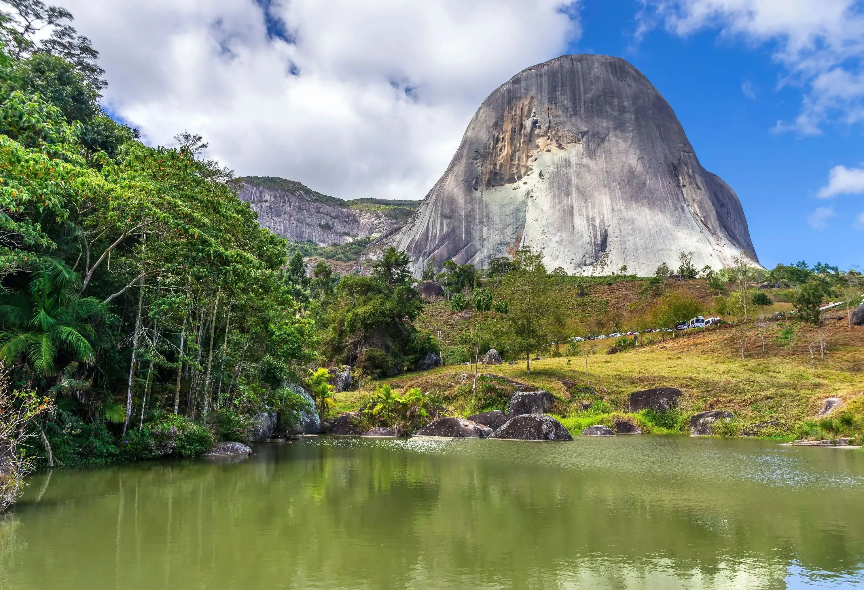 Rich vegetation surrounds a murky lake and a towering blue granite rock structure.