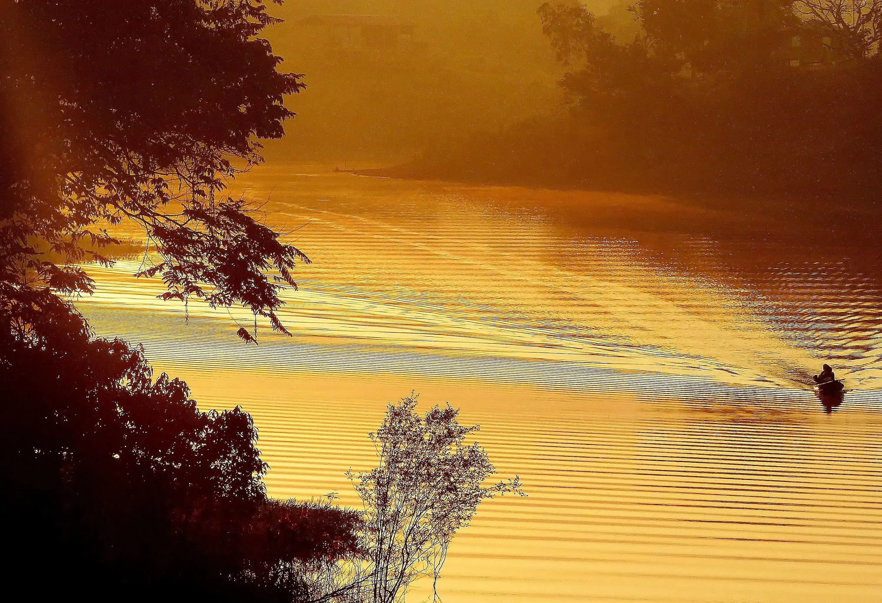 A man in a boat cruising across the rippling waters of a river stained by the golden hues of dawn.