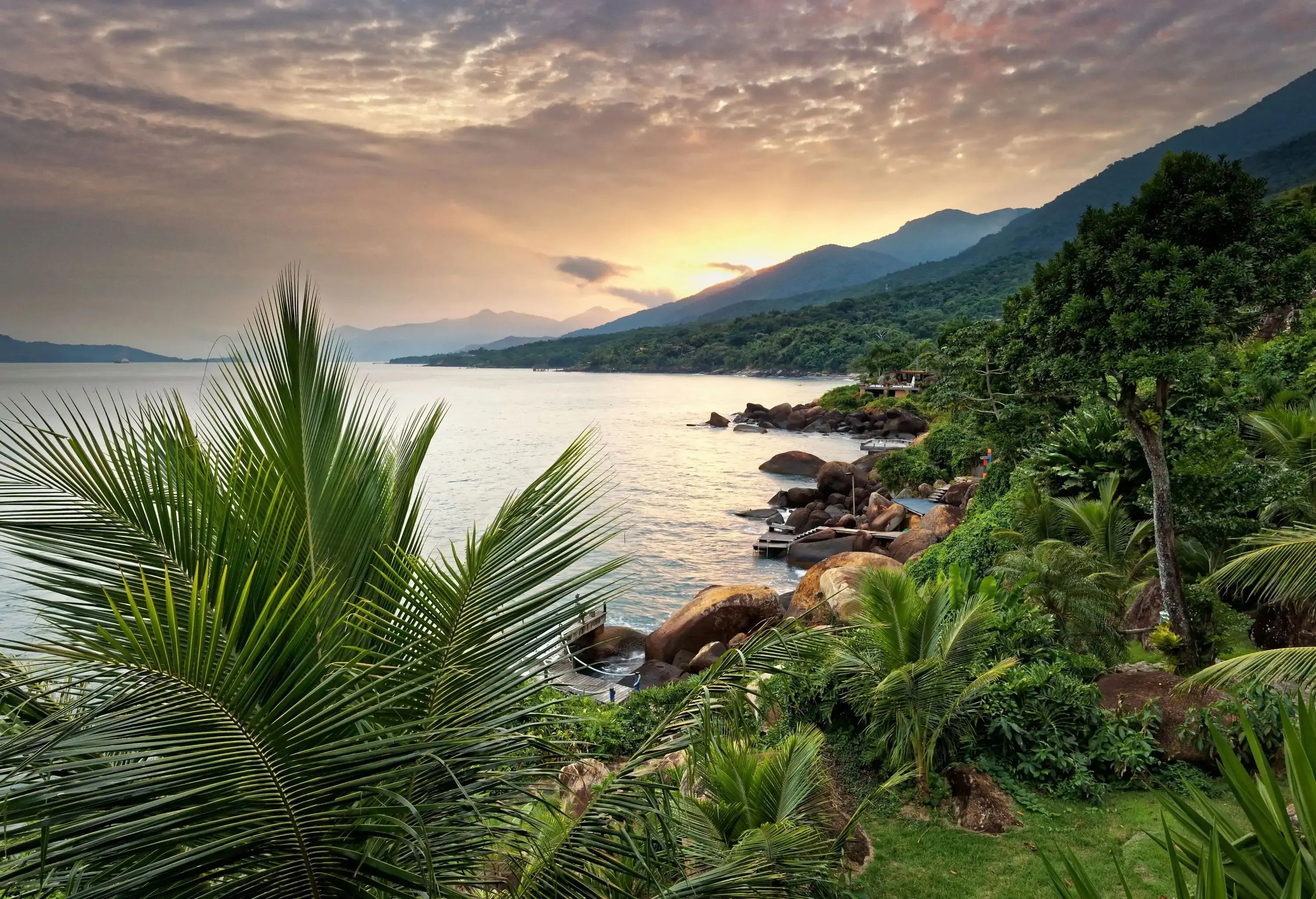 A scenic lush green landscape covers the coastline at the base of forested mountains against the dramatic sky.