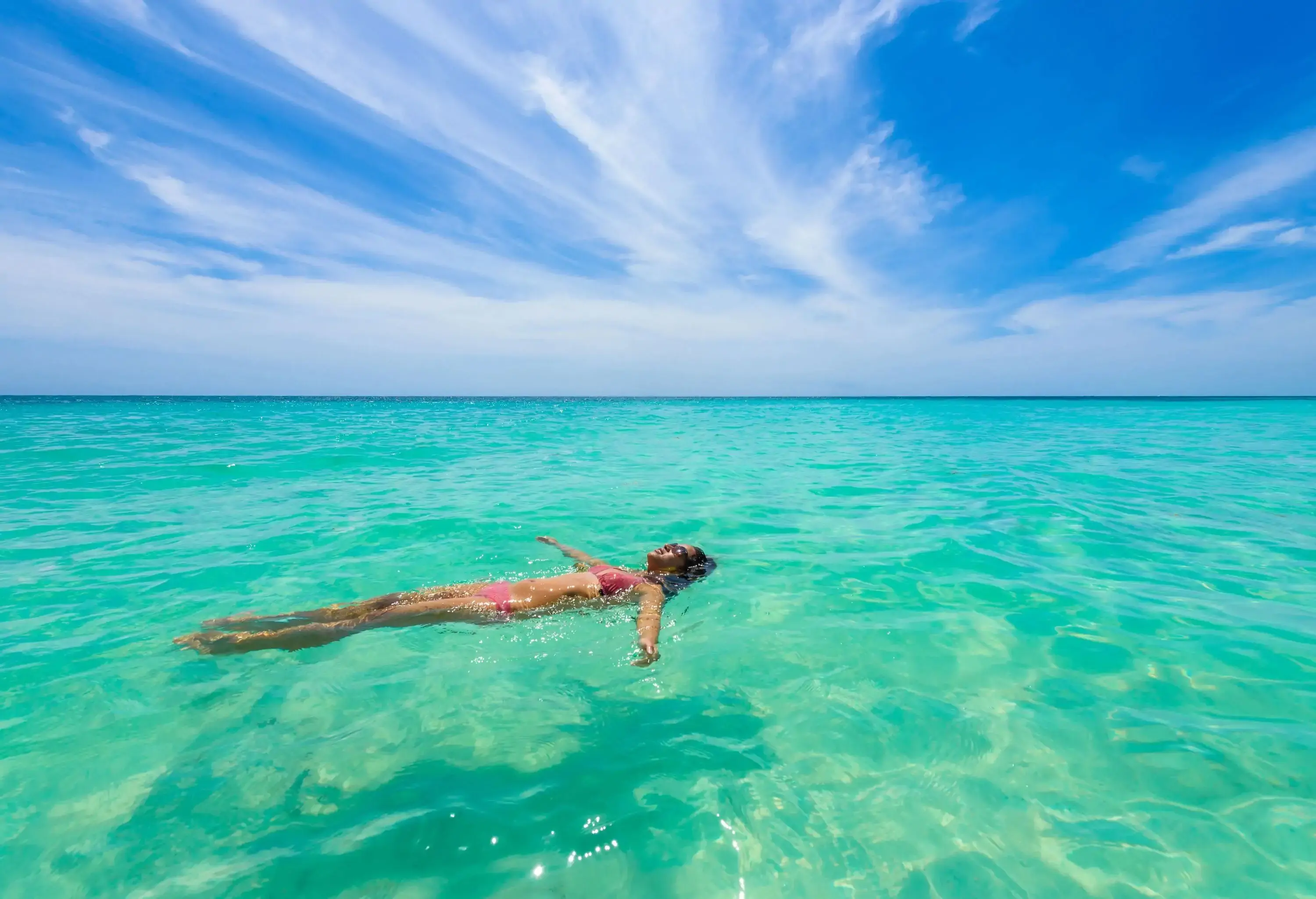 A carefree young woman in a bikini blissfully relaxes in the vibrant turquoise waters, effortlessly back-floating amidst the seemingly endless expanse of the vast sea.