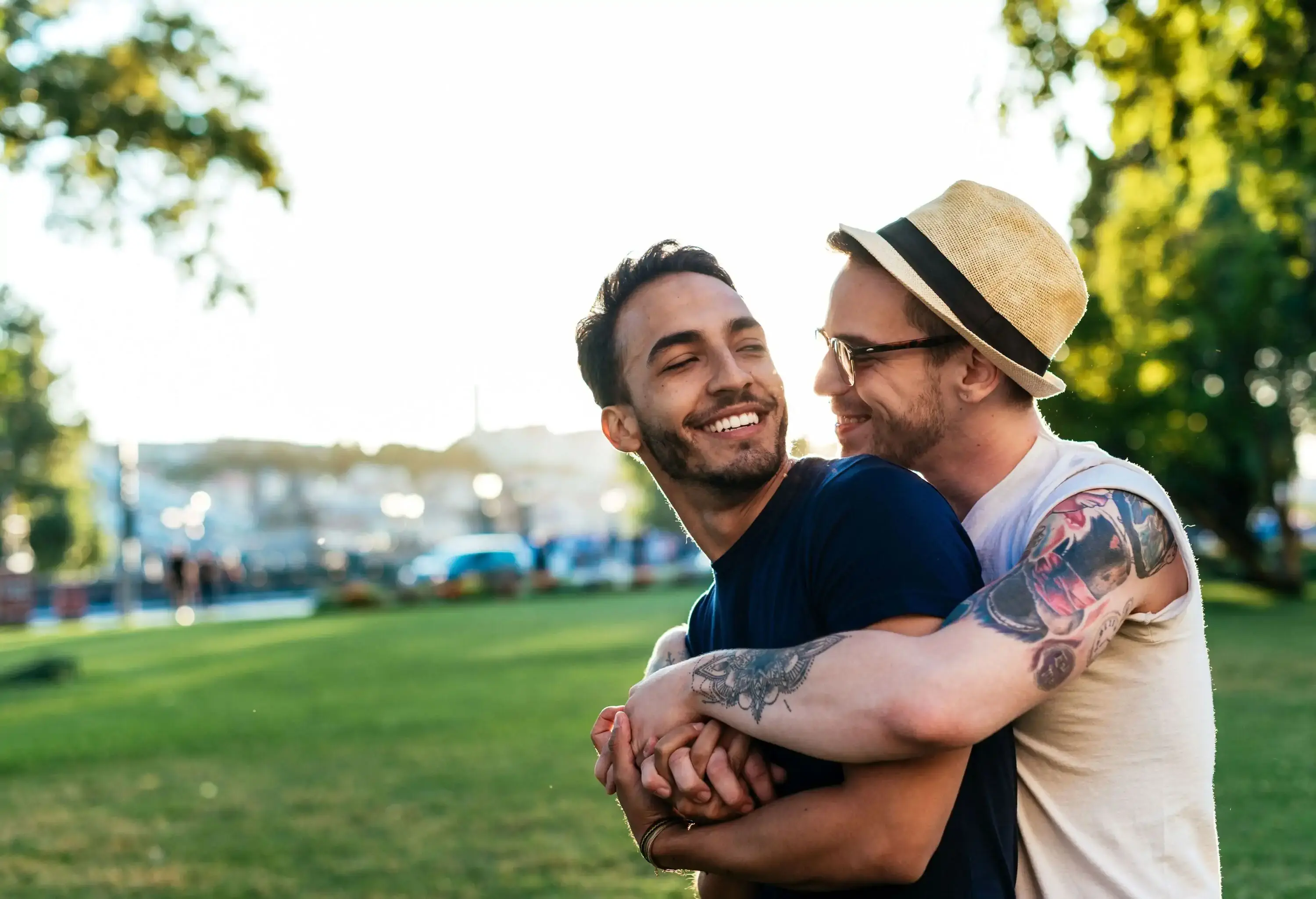 A tattooed gay man with a fedora hat and eye glasses hugging a male partner from behind as they smile and look into each other.