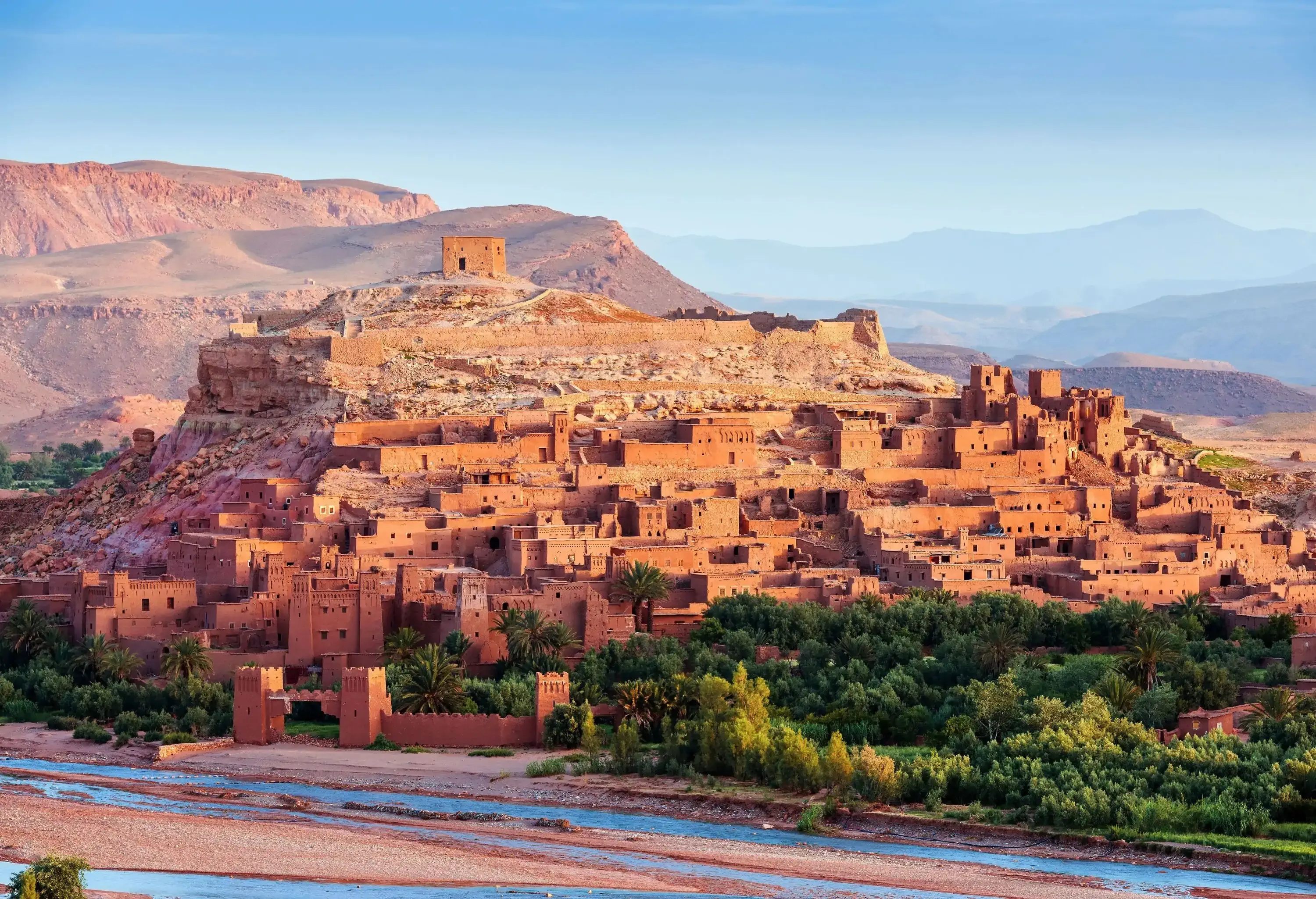 A fortified village with earthen buildings on the slopes of a hill next to a shallow river.