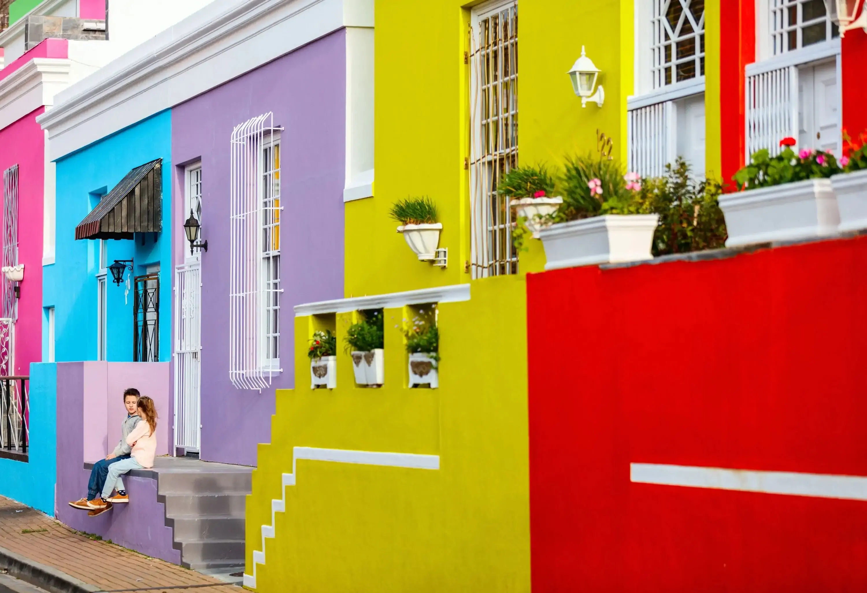 A boy and a girl kid sit at the doorstep of a row of vibrant, colourful houses.