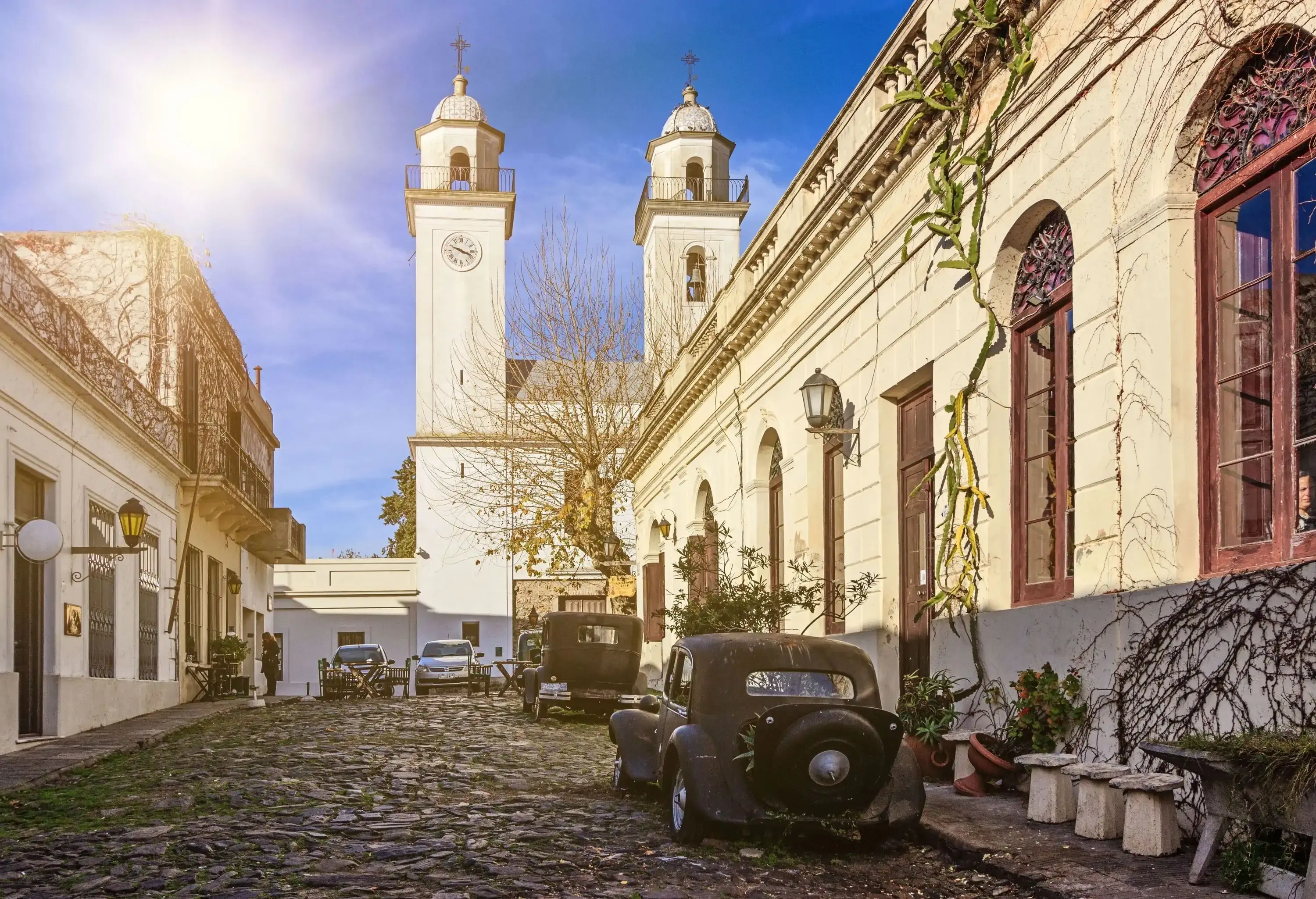 A cobblestone street with parked vintage cars along colonial buildings with views of a twin belfry in the background.