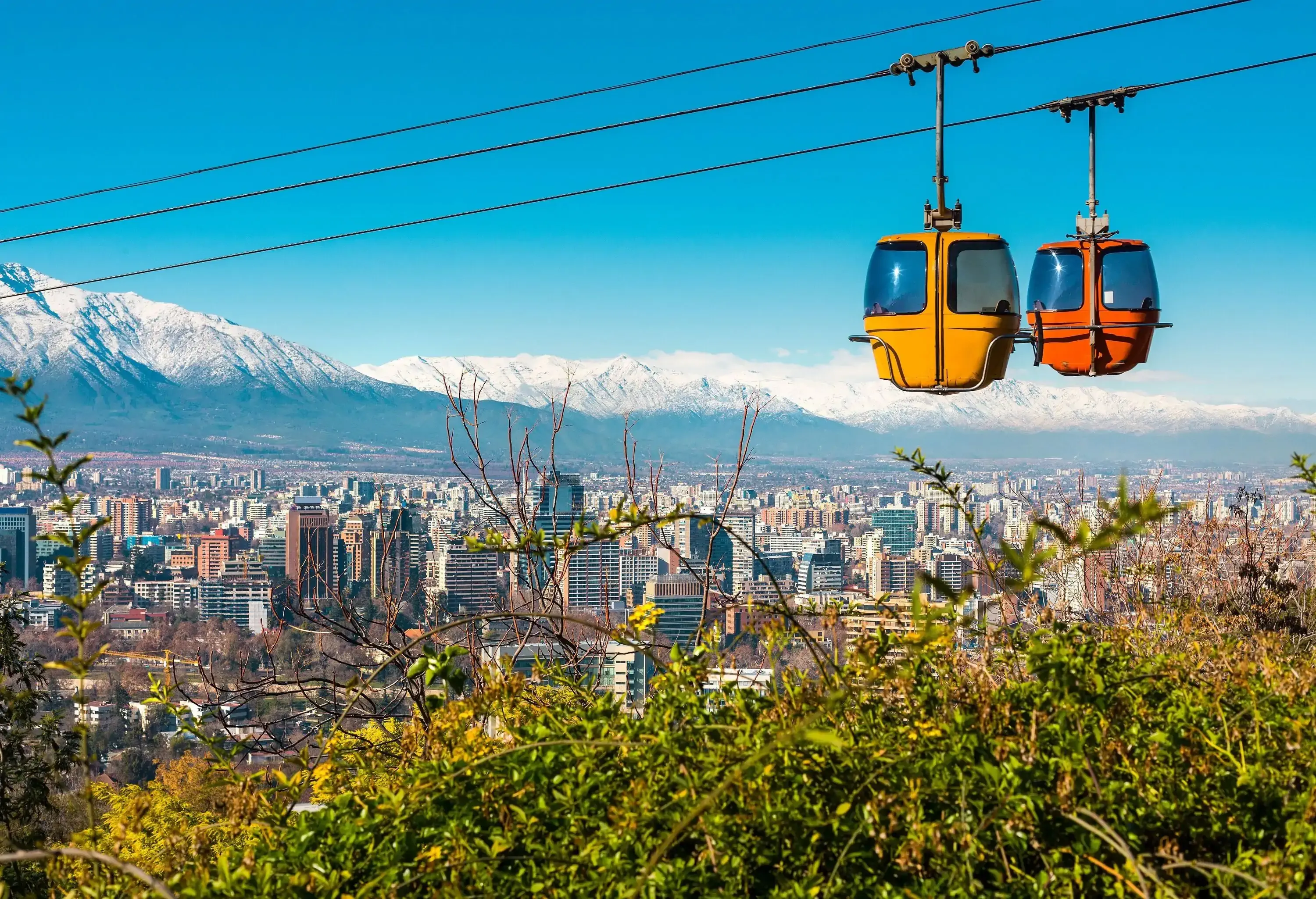Two cable cars travelling on a cable against the backdrop of a cityscape and snow-capped mountains in the distance.