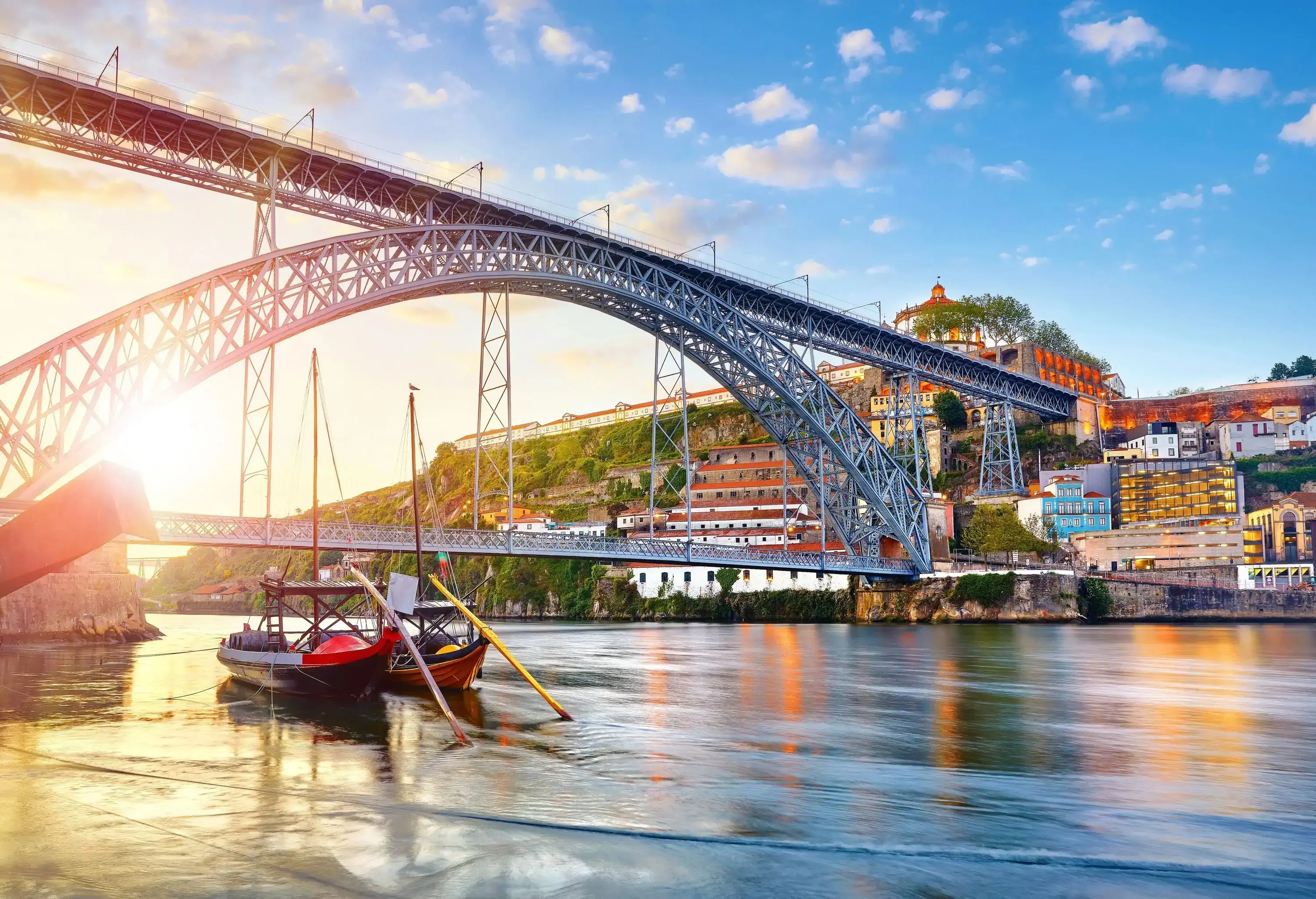 Two boats travel under a metal arch railroad bridge across a calm river along the waterfront buildings.