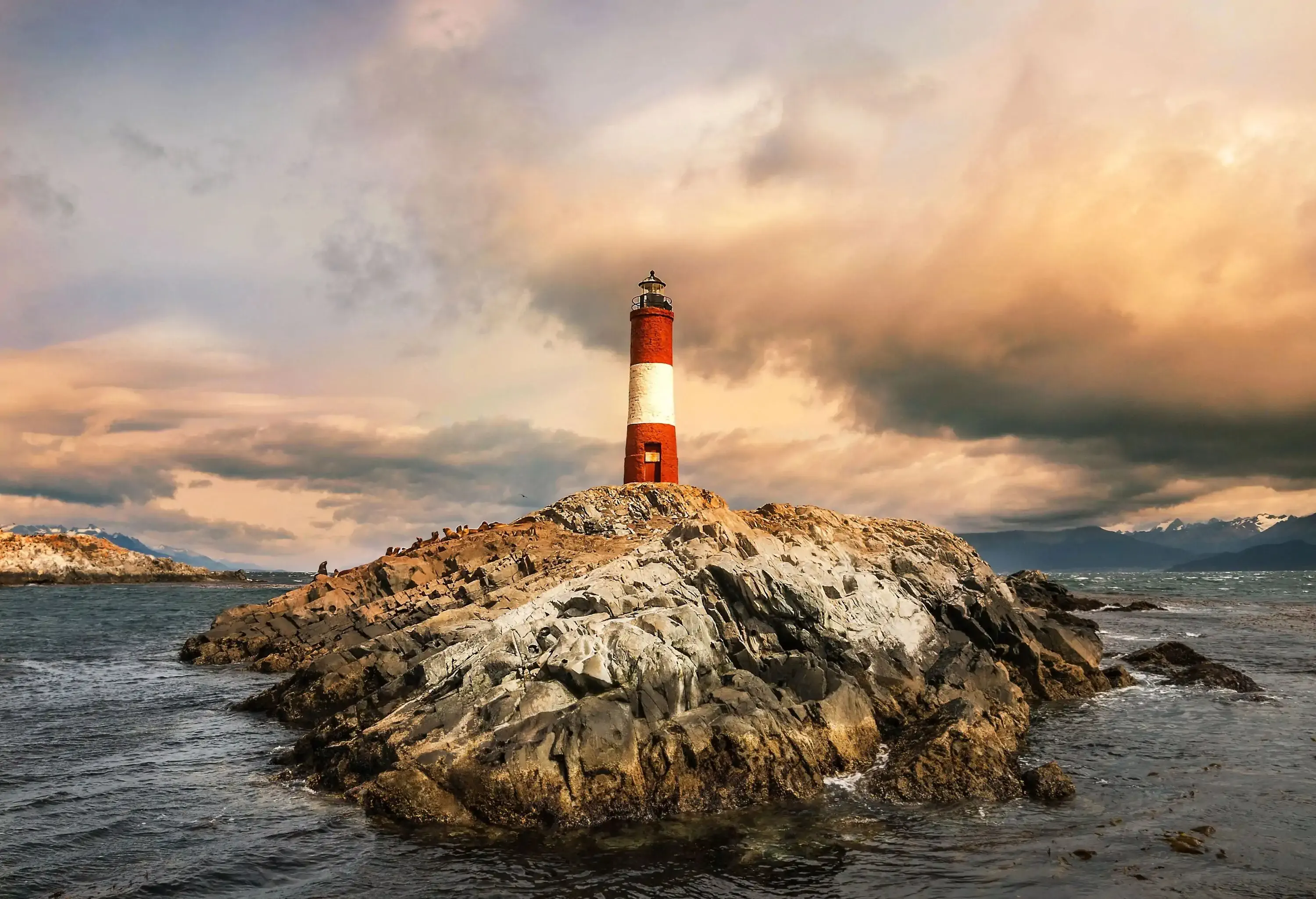 A red with a broad white band cylindrical lighthouse topped with a black lantern emerges from a rocky outcrop above the sea.