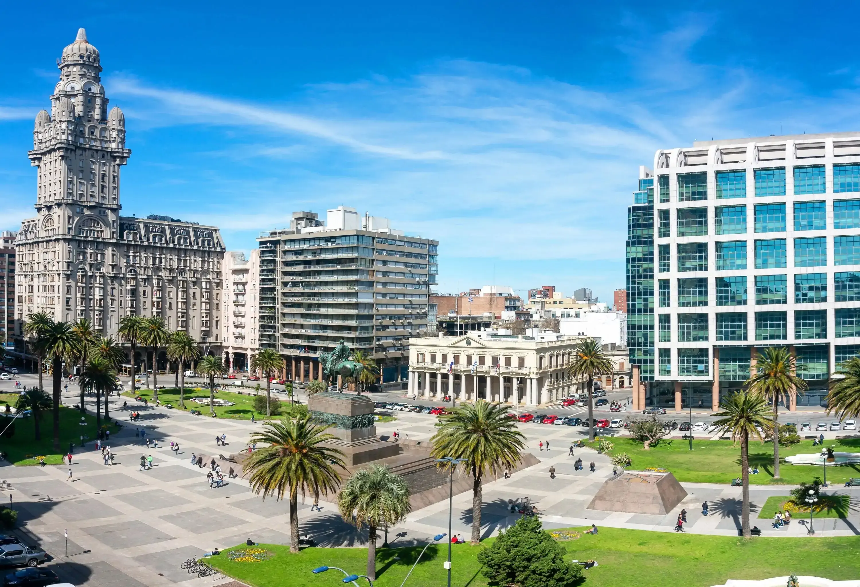 Several people are passing by the city square dotted with trees and a bronze equestrian statue surrounded by commercial buildings.