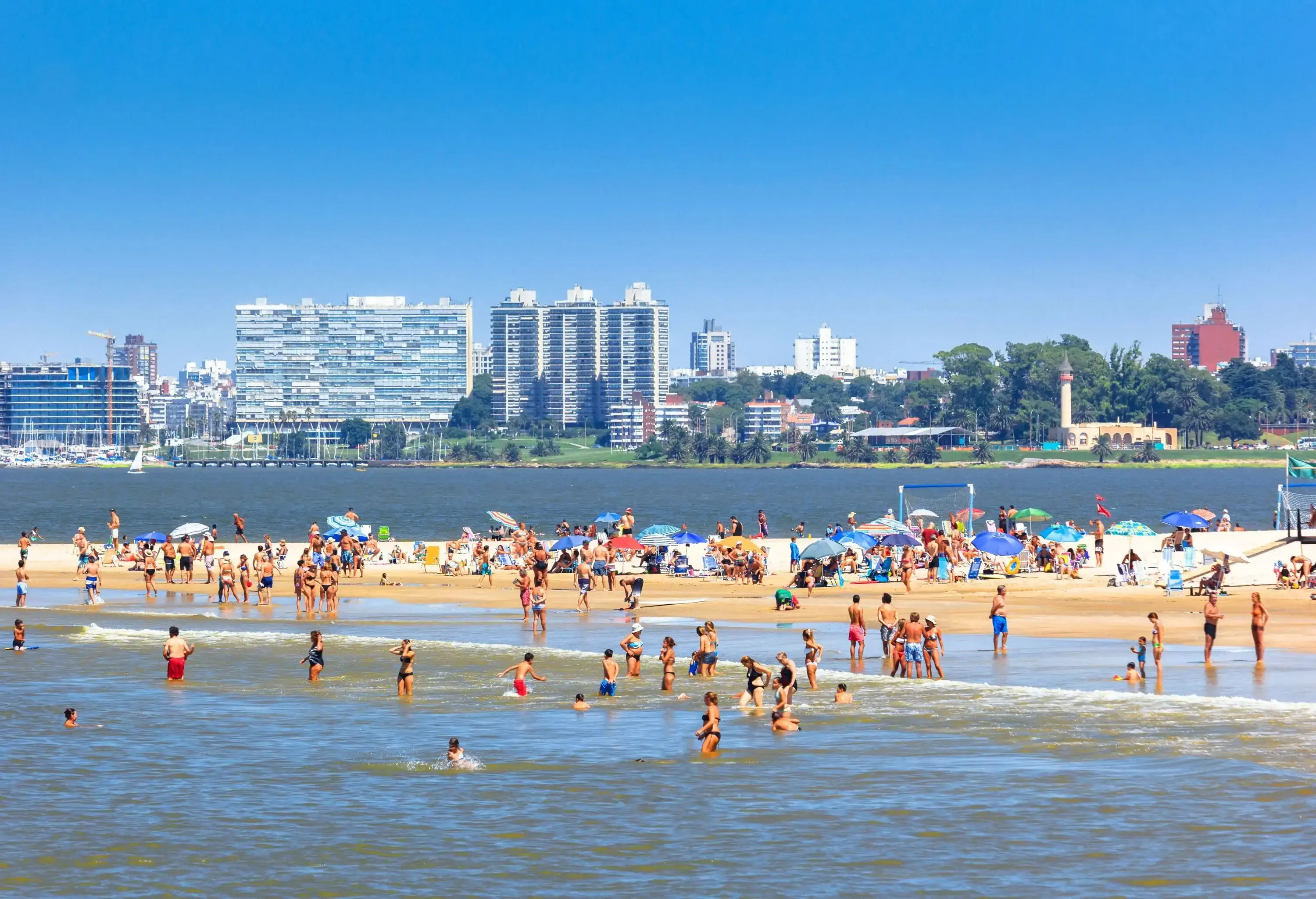 Beachgoers enjoy the soft sands and the calm waters on a sunny day against the skyline in the background.