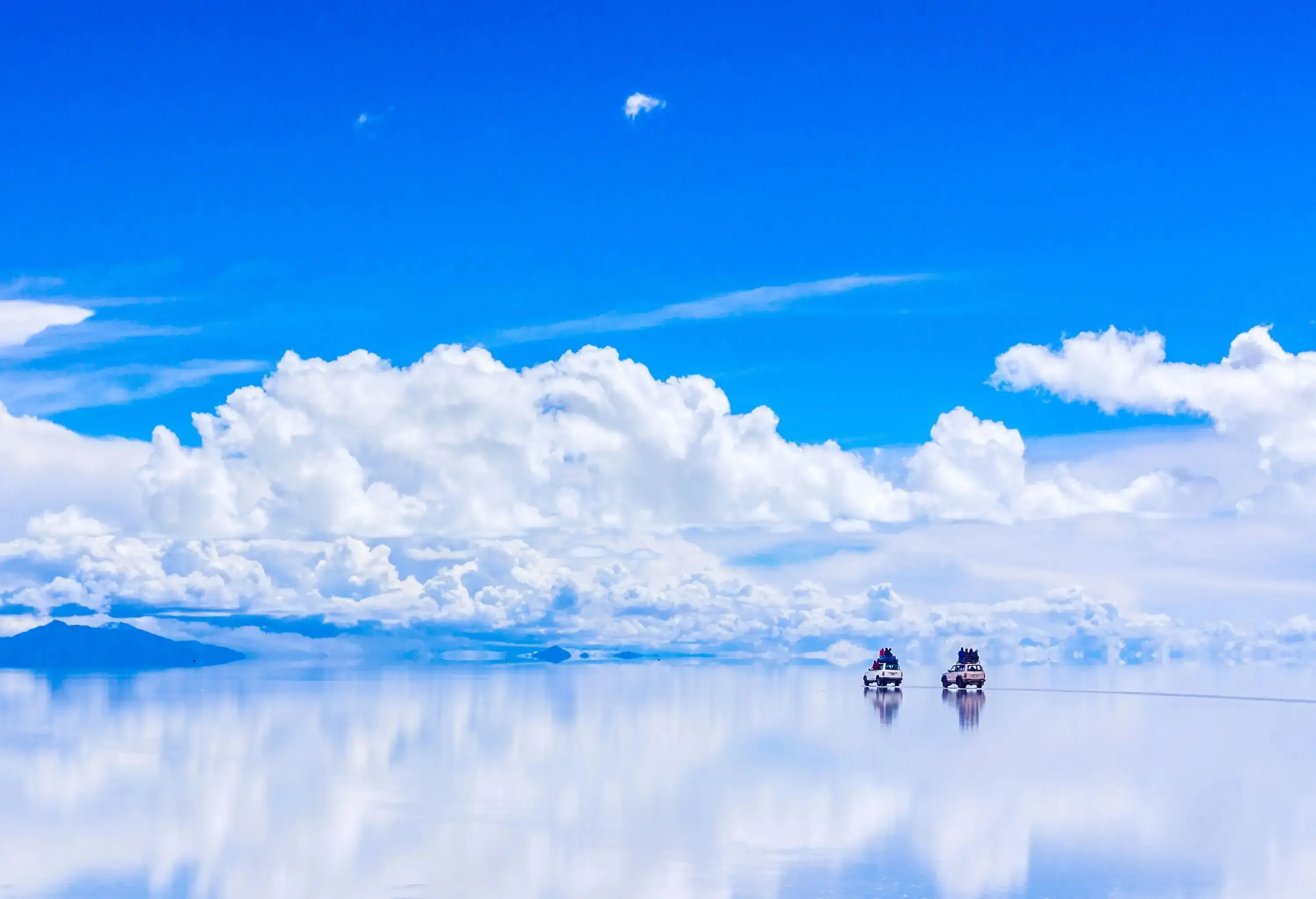 Two SUVs moving over the shiny white surface of the salt flats, with puffy clouds floating in the sky.