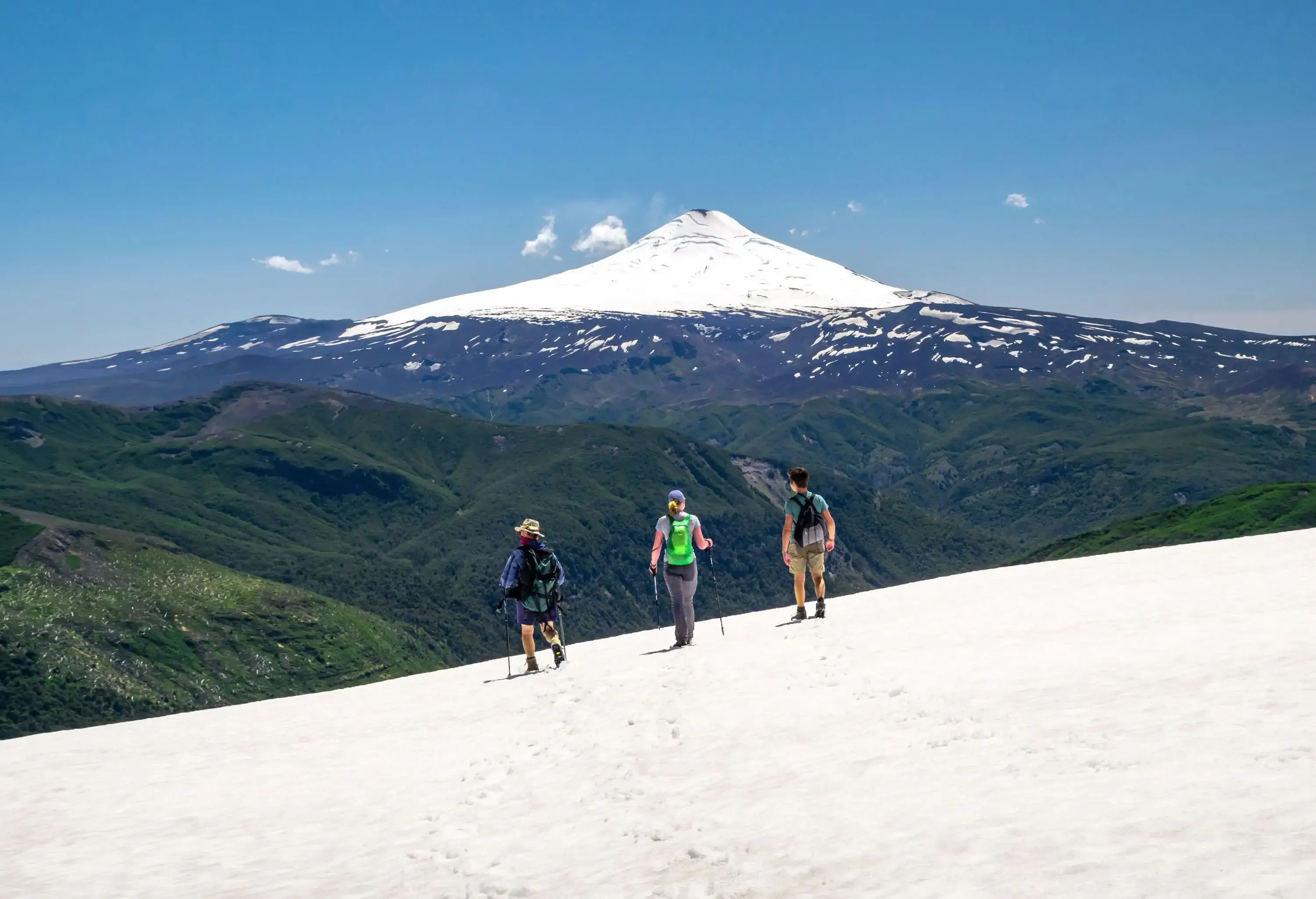 Three people hike through a white snowy land overlooking the forested mountains and the snowcapped volcano against the clear blue sky.