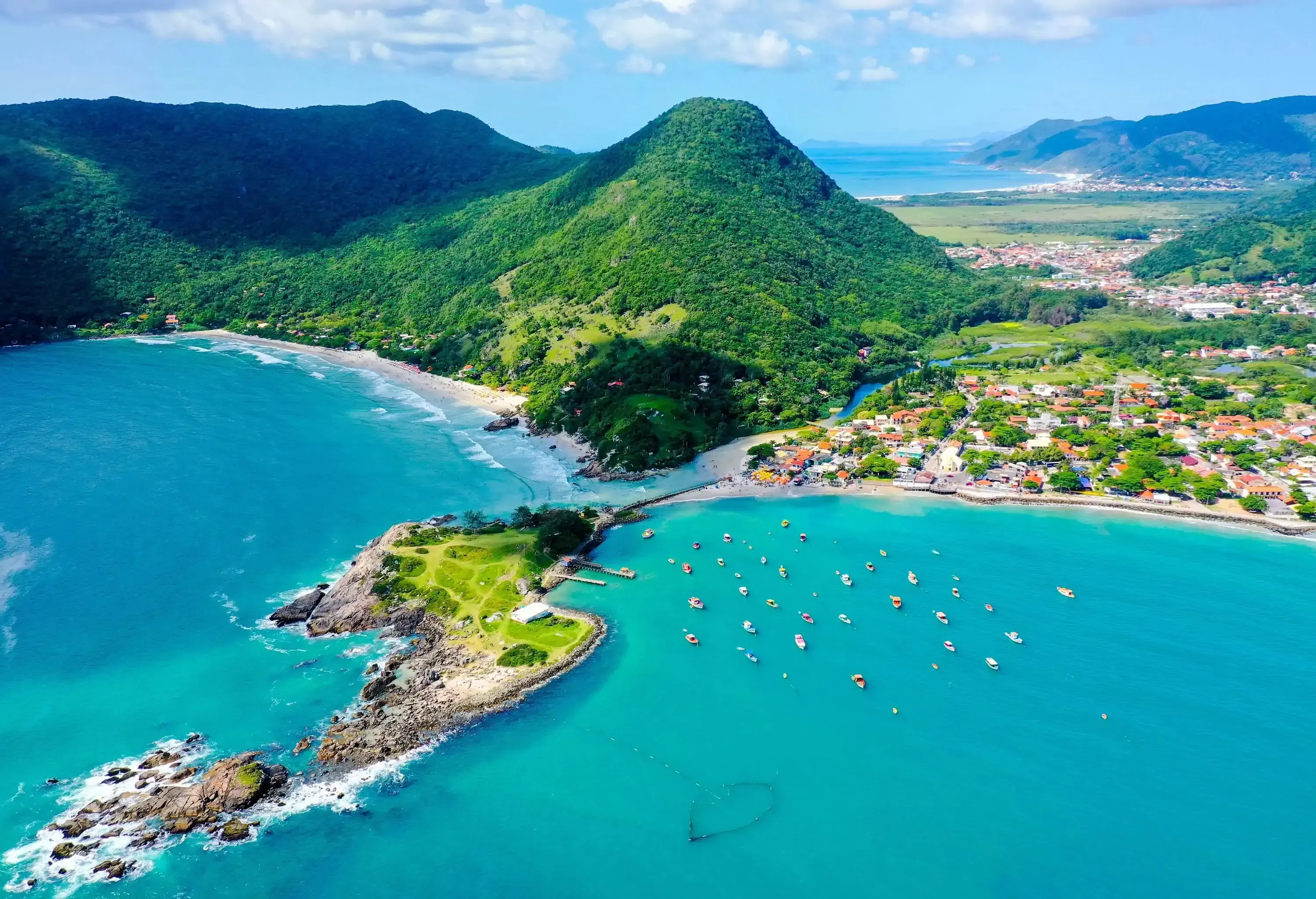 A charming town nestled at the foot of green-covered hills, with a serene beach and boats moored near the shore adding to its coastal allure.