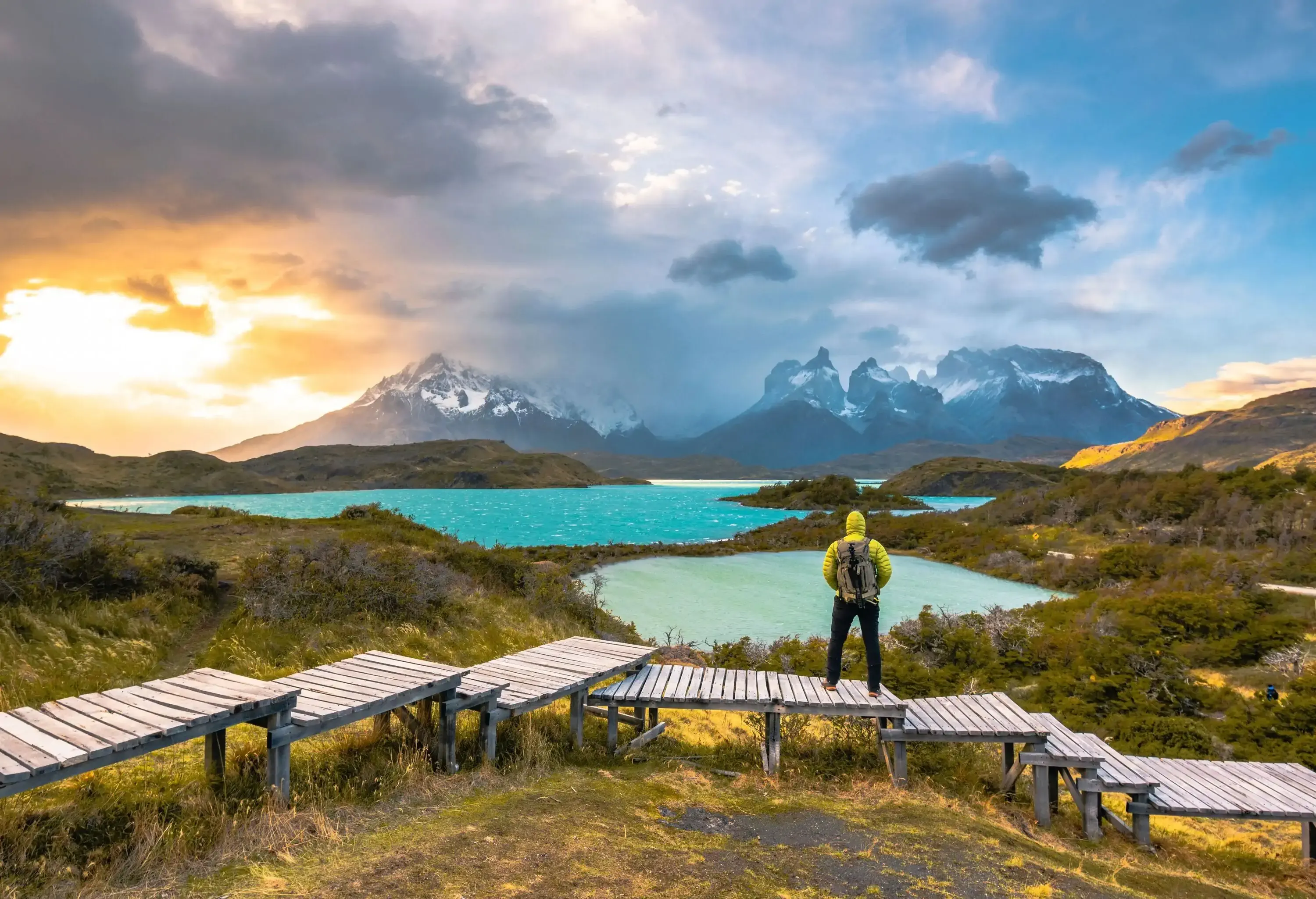 A backpacker standing on a tiered wooden boardwalk overlooking a lake below the mountains.