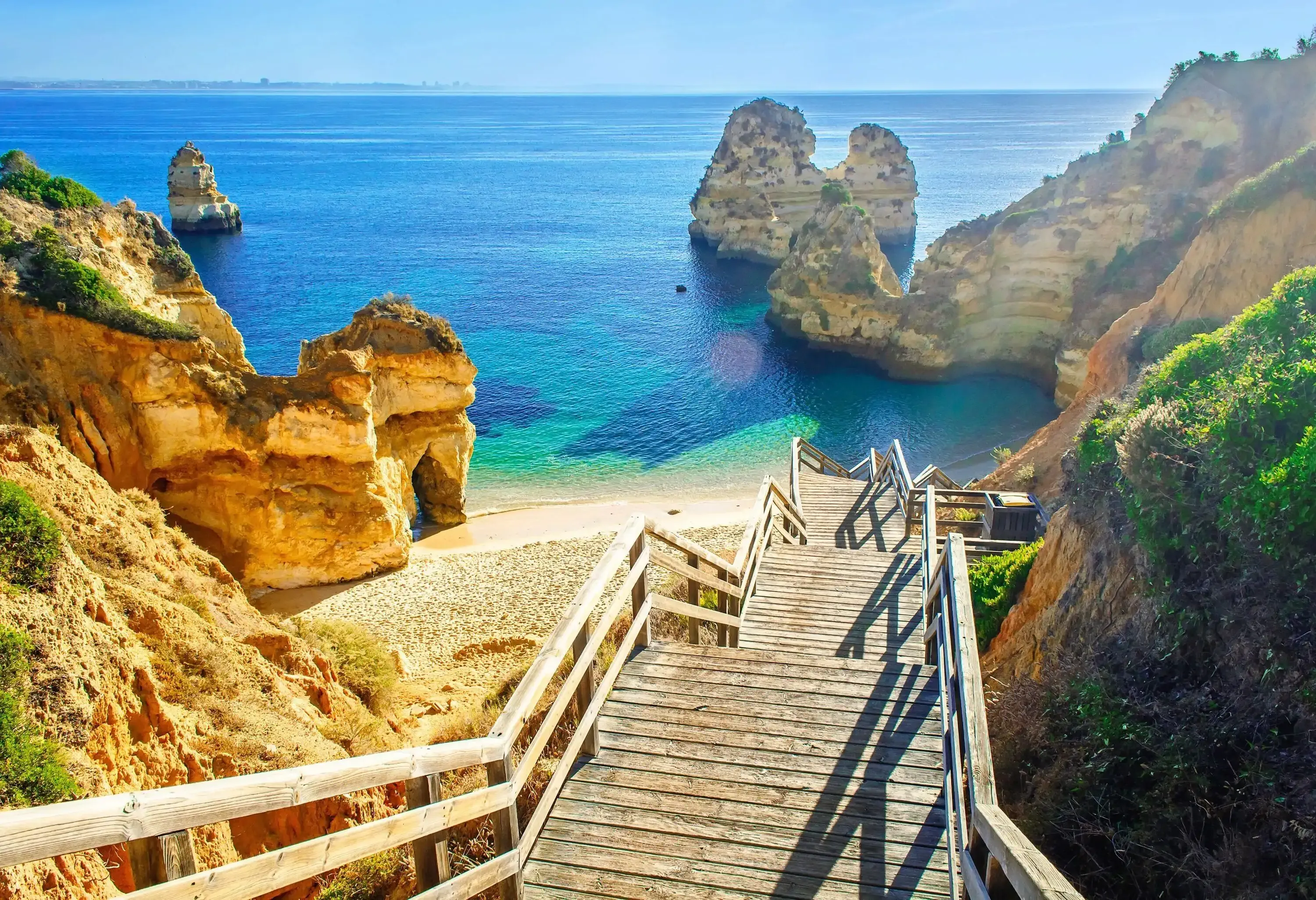 A wooden staircase leads to a beach surrounded by natural rock formations with lush cliffs and calm blue waters.