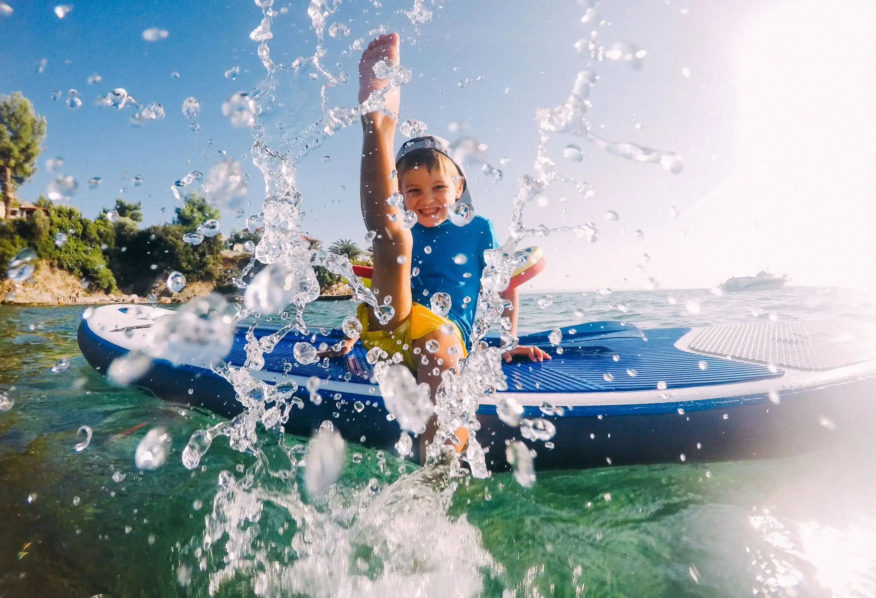 A lively little boy sits on a stand-up paddleboard, gleefully splashing water with his feet.