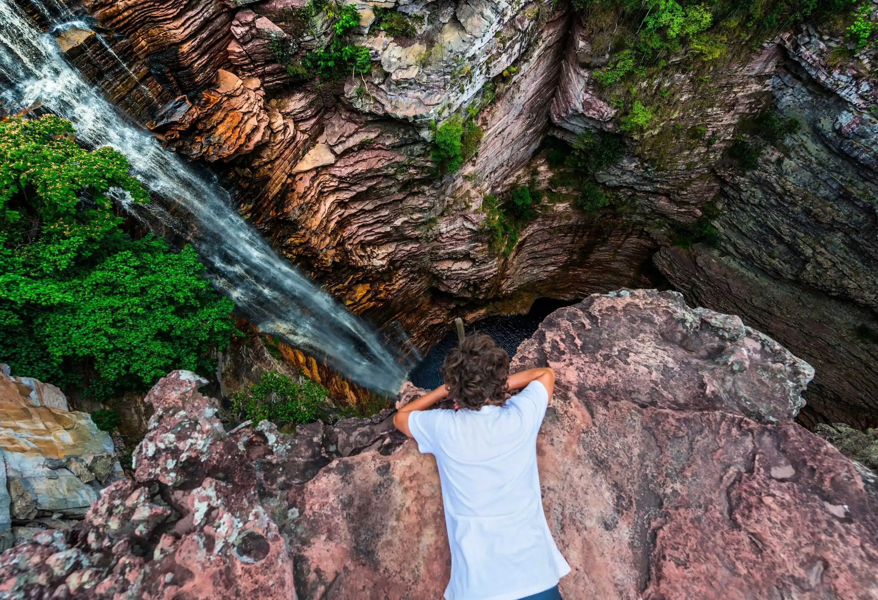 A man in a white shirt lies on a cliff's edge and looks down at a cascading waterfall on a steep cliff.