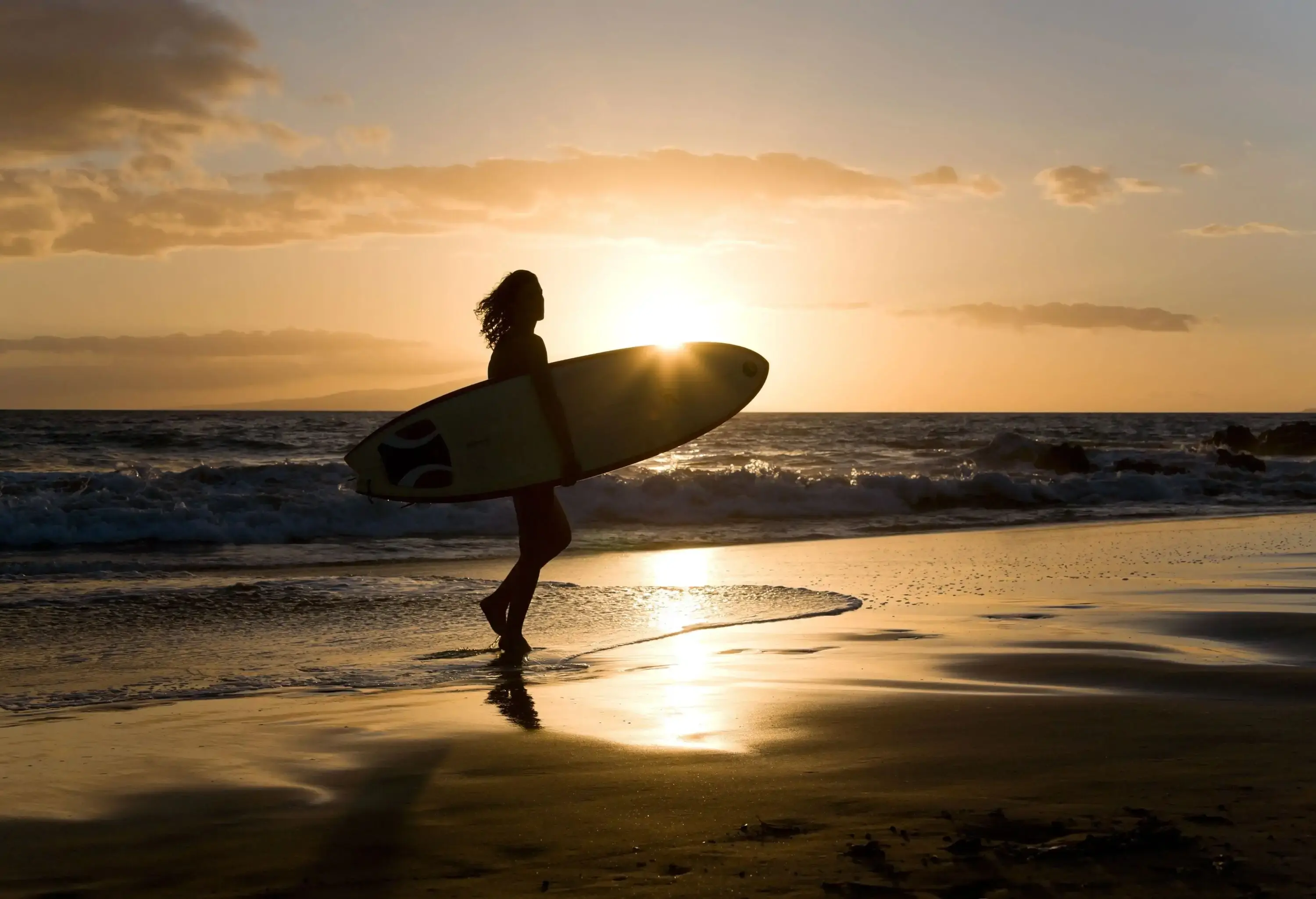Silhouette of a woman with a surfboard on the shore of the beach at sunset.