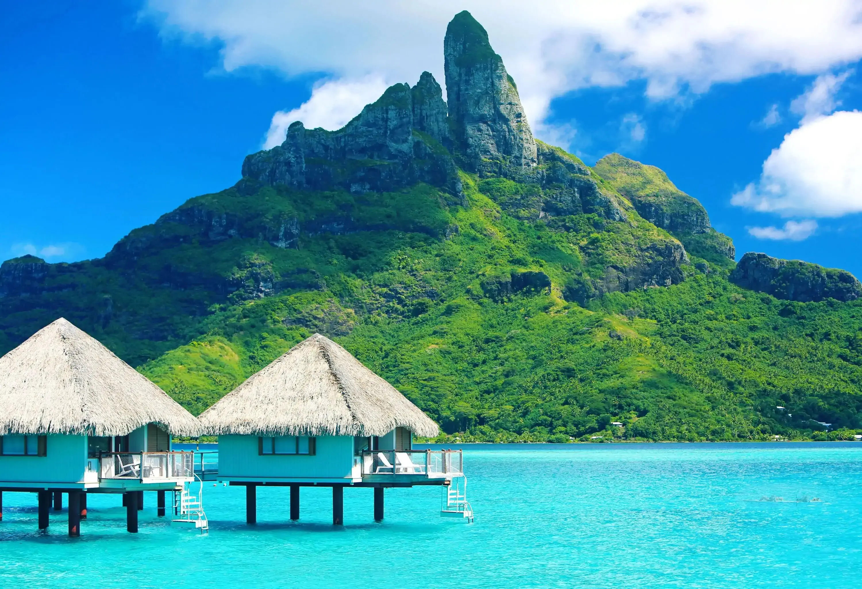 Luxurious nipa huts over the azure sea overlooking a forested and rocky mountain against the cloudy blue sky.
