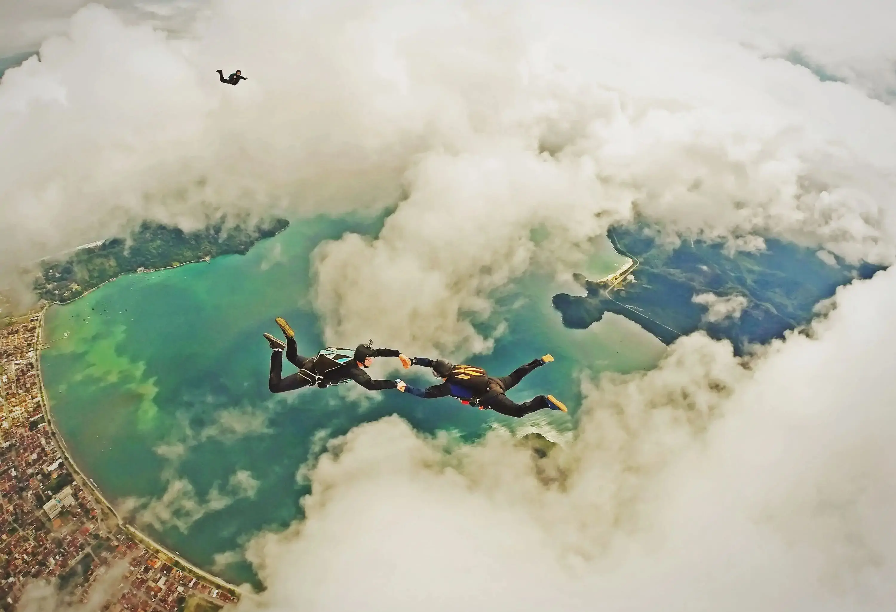 Two people hold each other's hand as they skydive above a cityscape, a bay, and thick clouds.