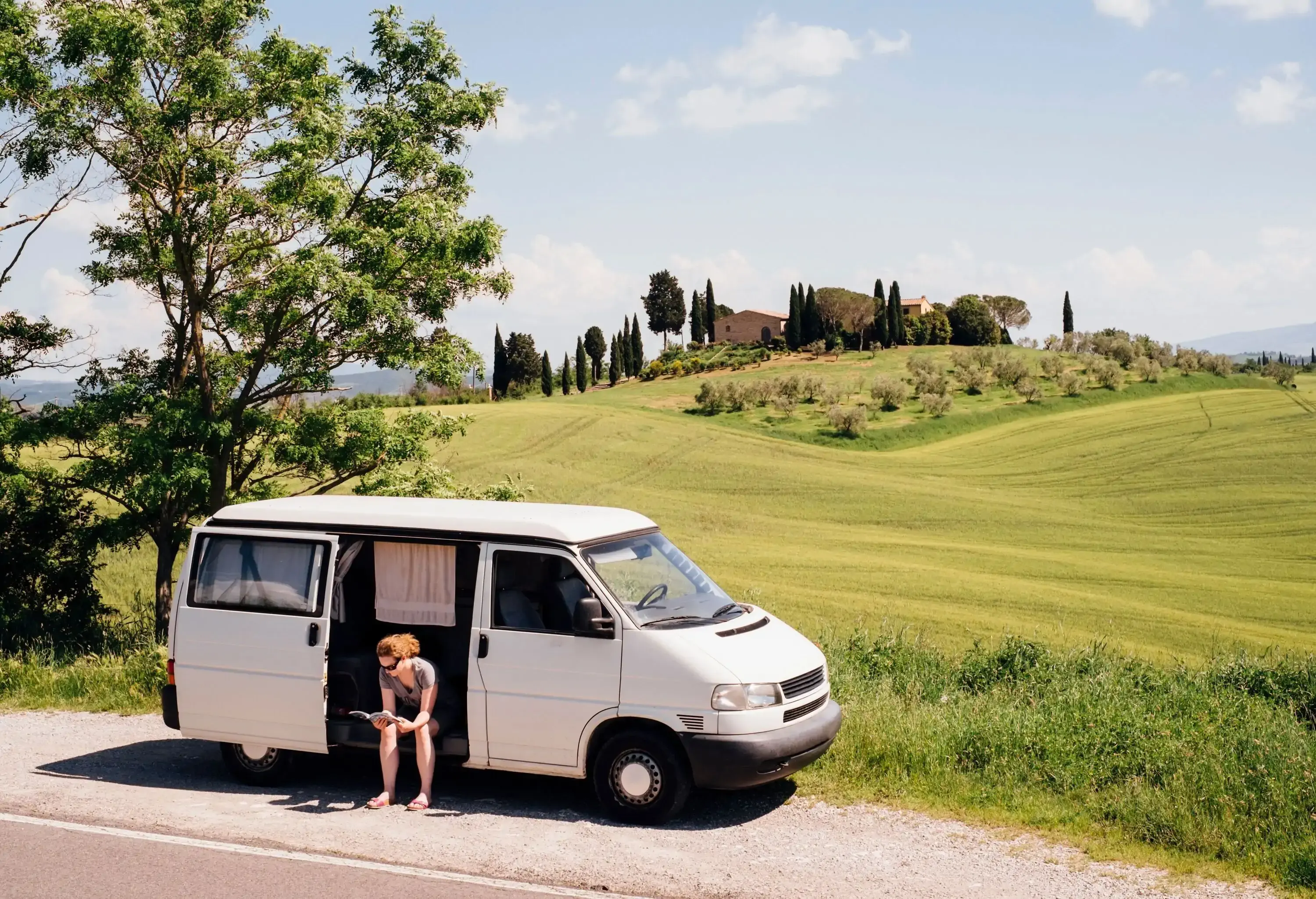 A woman in sunglasses sits at the side of a van parked along a grassland, reading a book.