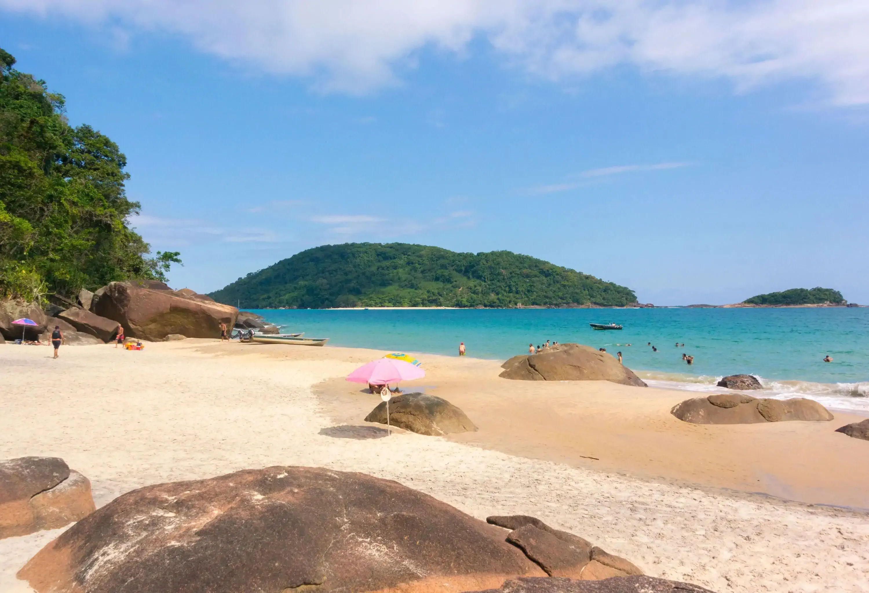 Boulders on the sandy shore of the turquoise sea dotted with beachgoers.