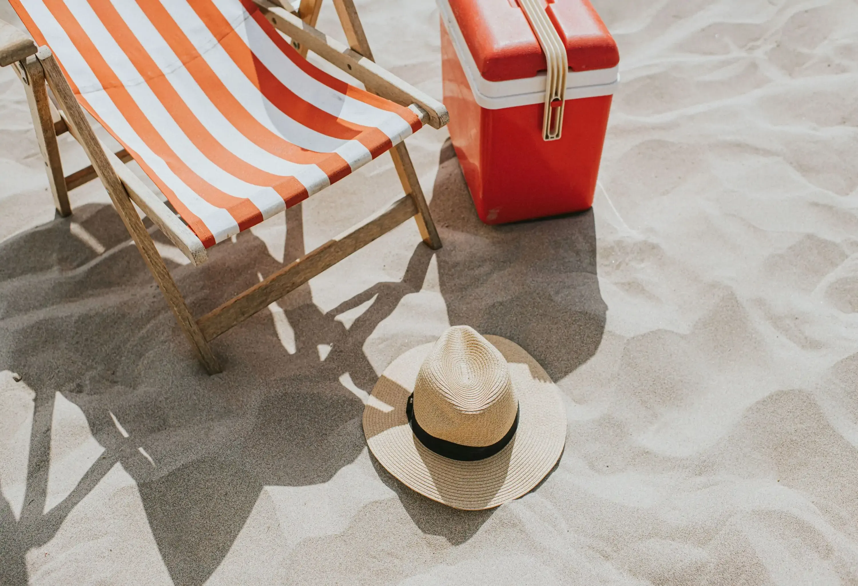 A relaxing coastal scene. A traditional style striped deckchair sits on a sandy beach. A sun hat rests on the ground and a cool box / cooler sits off to the side. Sand provides space for copy.
