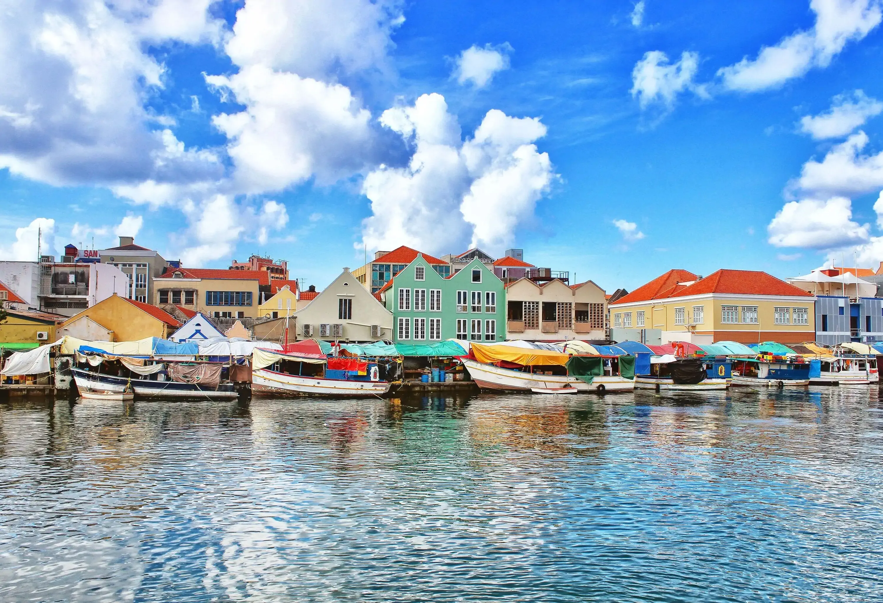 A lively promenade adorned with colourful boats, brimming with merchandise, gracefully moors along the waterfront, while buildings provide a charming backdrop.